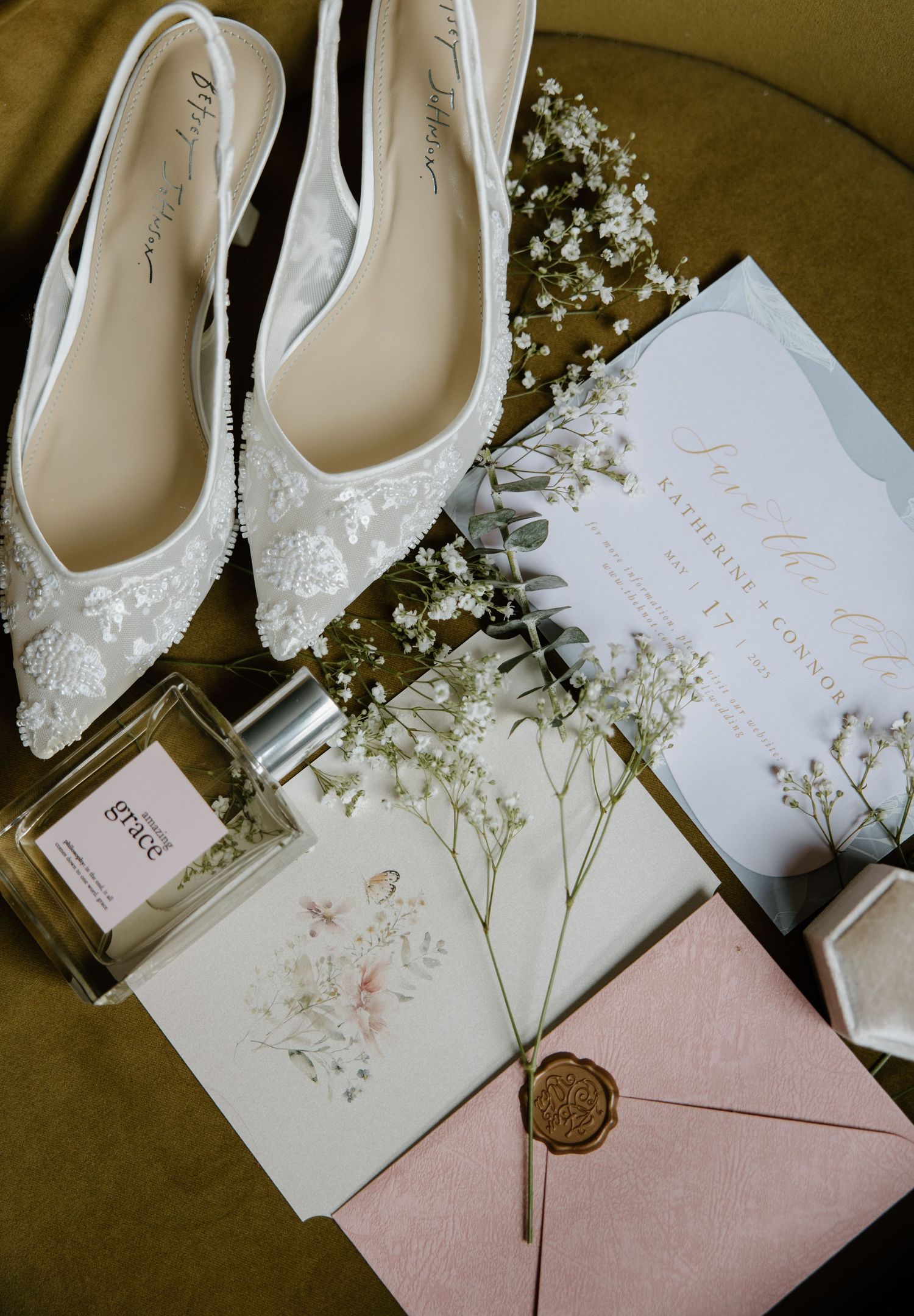 White lace bridal shoes arranged with wedding invitations, baby's breath flowers, and perfume bottle on olive velvet surface.