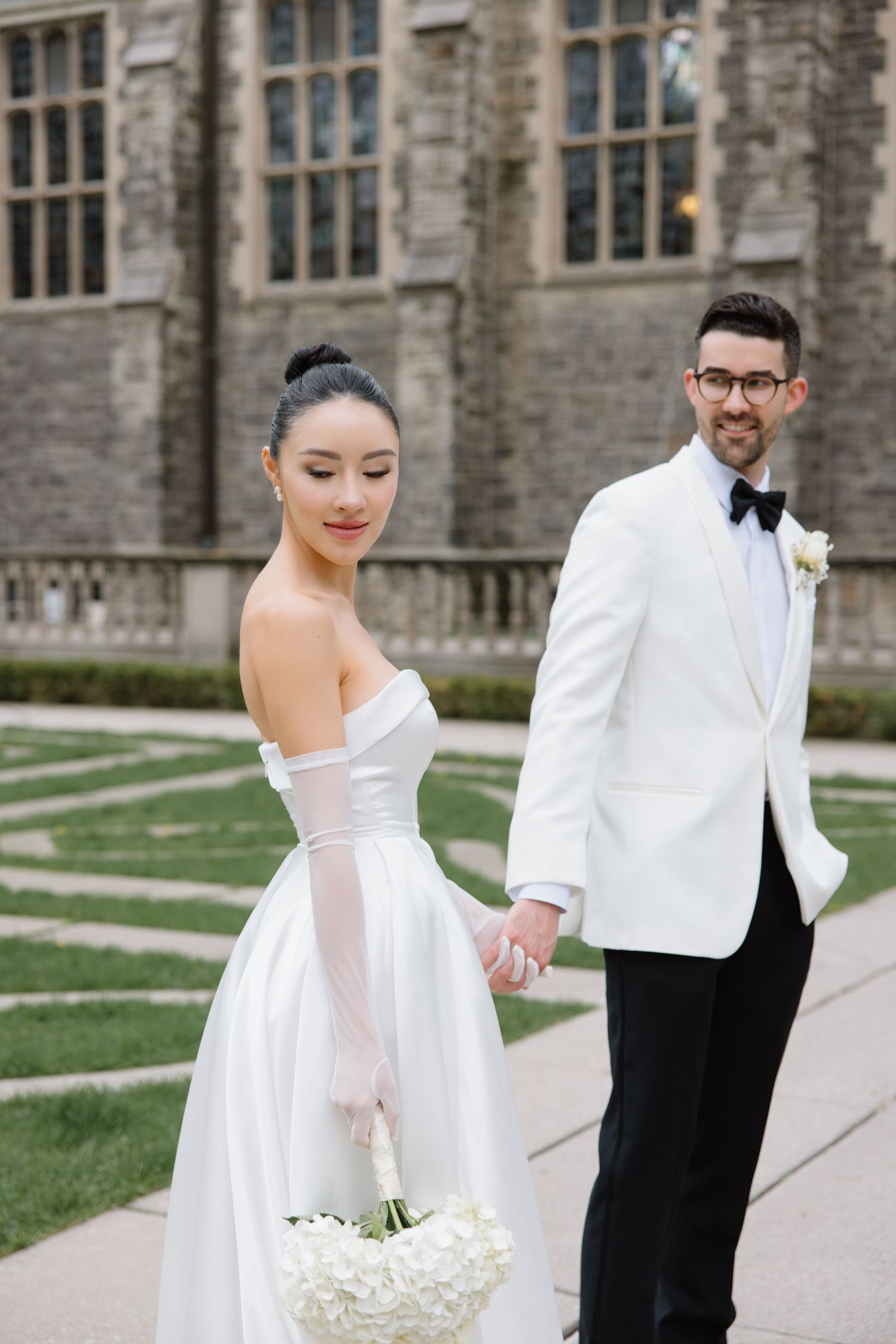Elegant couple in formal wedding attire holding hands outside a gothic stone building with a white bouquet.
