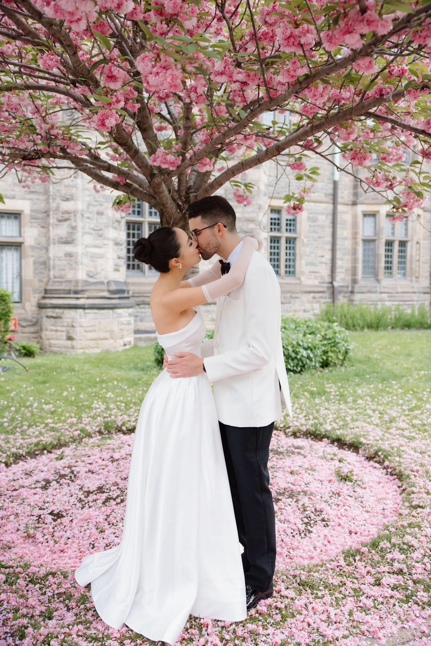 A romantic couple in wedding attire dancing under blooming cherry blossoms with falling pink petals and stone building.