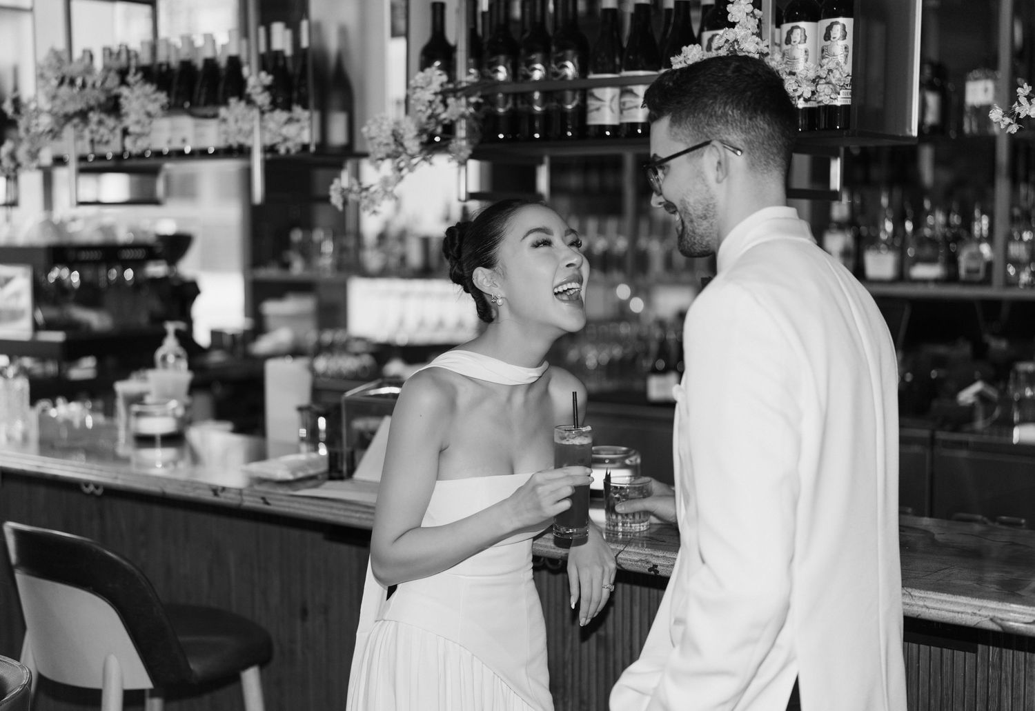 A couple in formal attire share a joyful moment while having drinks at a vintage bar counter.
