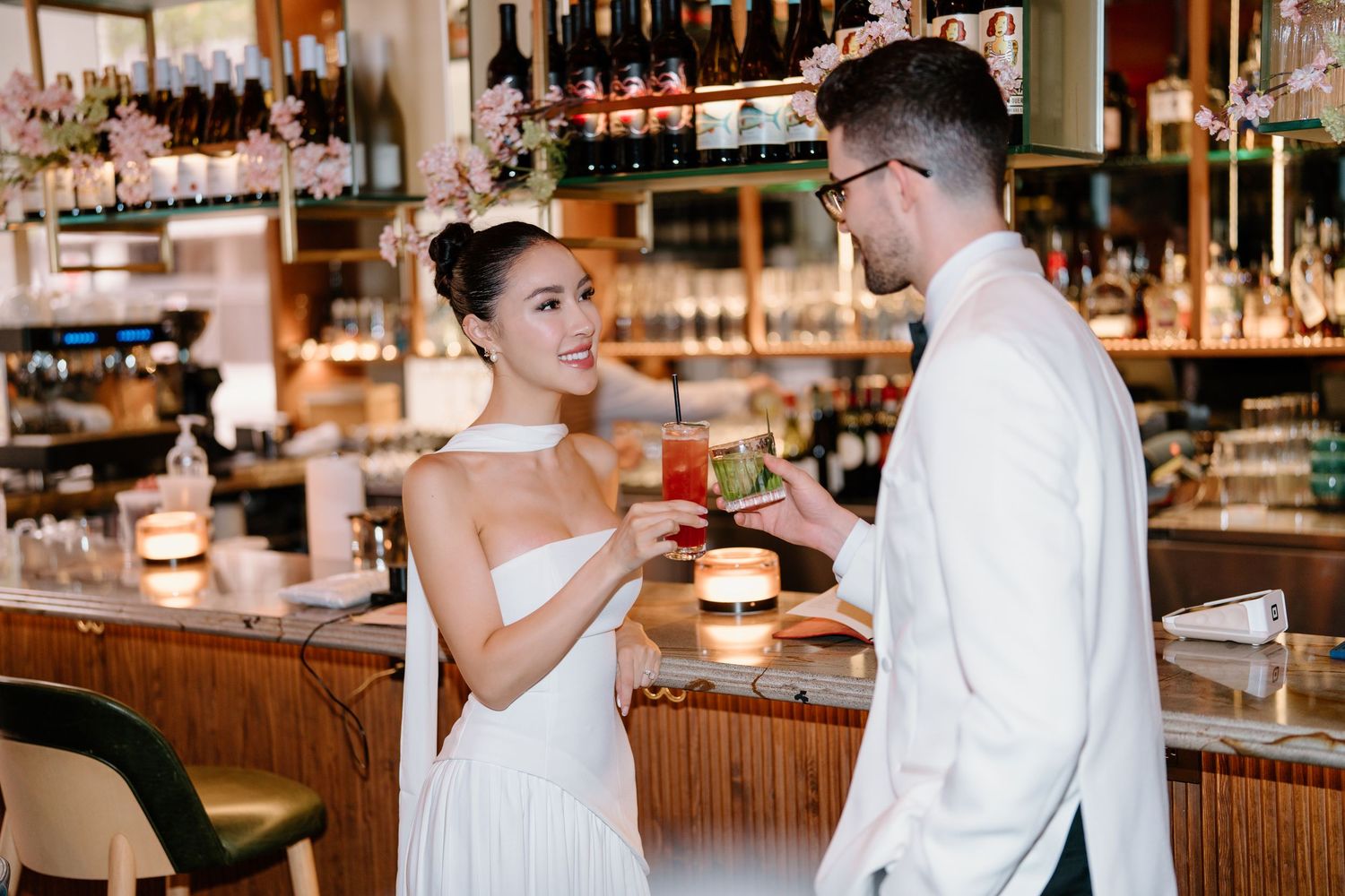 Two people in white outfits share drinks at an elegant bar counter with wine glasses and bottles displayed behind them.