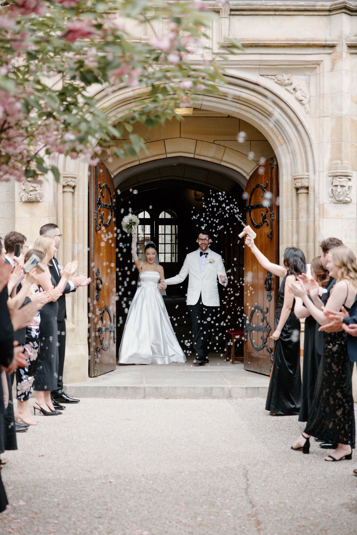 Wedding guests celebrate as newlyweds exit historic stone archway entrance with confetti falling amid elegant floral decor.