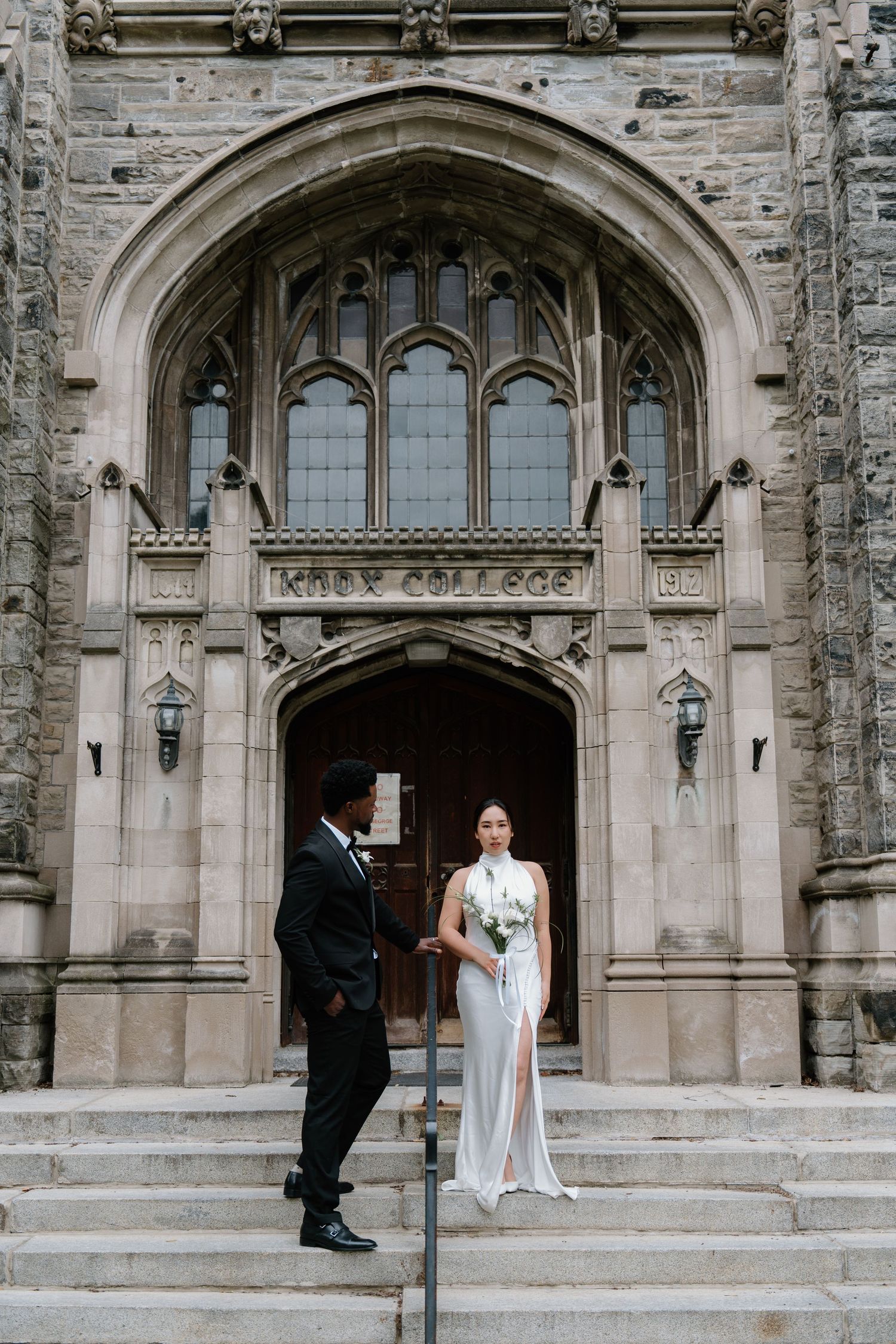 Wedding couple stands on stone steps in front of a grand Gothic style church entrance with arched doorway and lanterns.