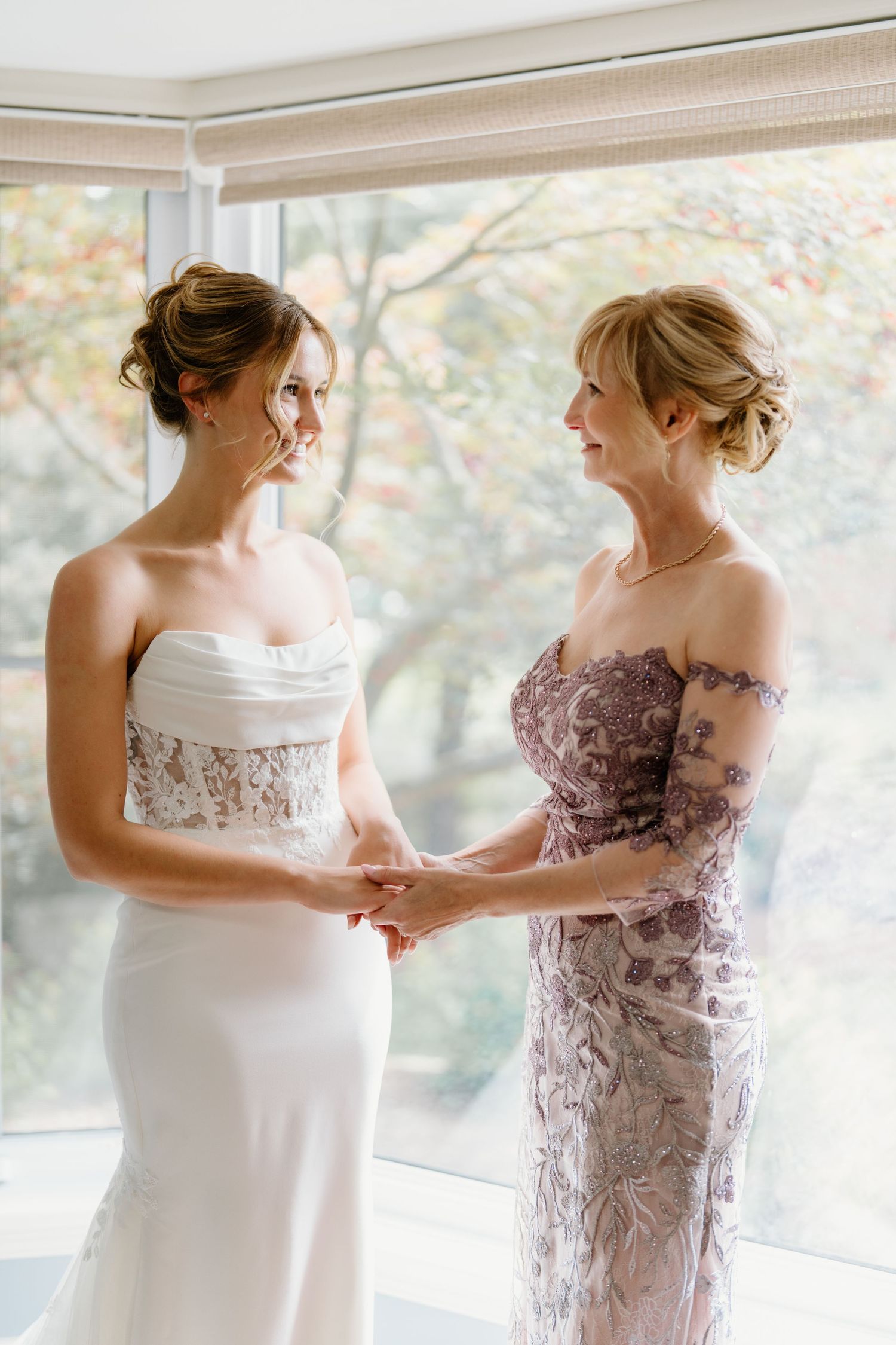 Two women in elegant dresses share a tender moment by a window, one in white and one in mauve lace.
