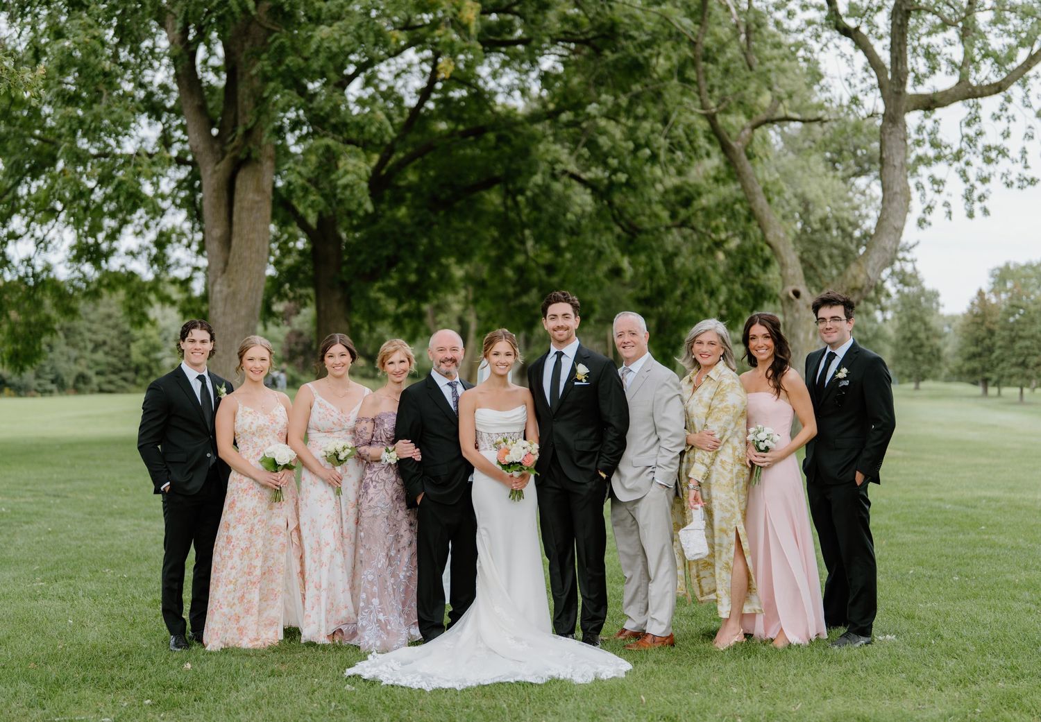 Wedding party poses together on a green lawn with trees in the background.