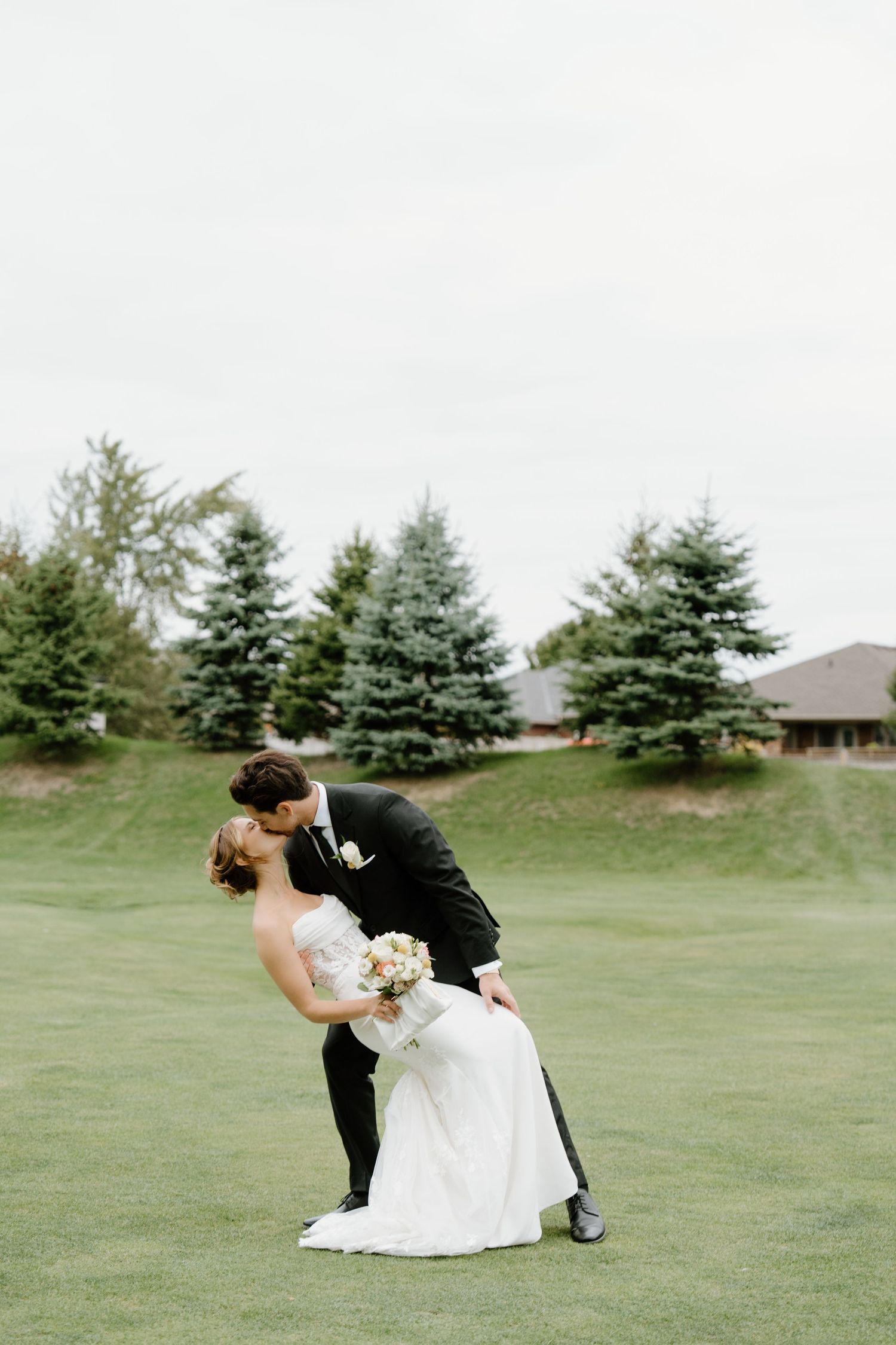 A romantic dip pose between newlyweds on a grassy lawn with evergreen trees in the background.