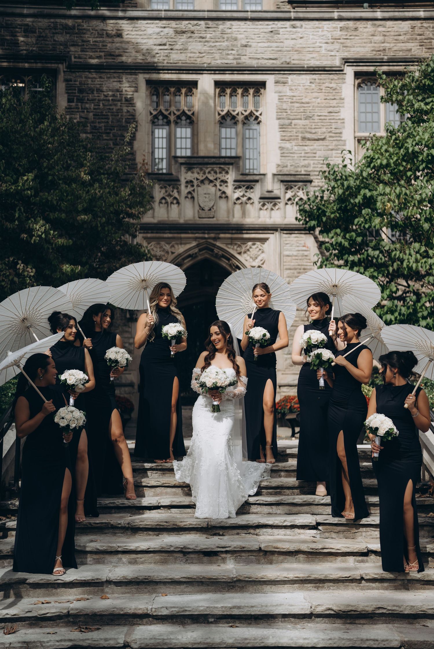 Wedding party poses with white umbrellas and bouquets against stone building backdrop in classic black and white attire.