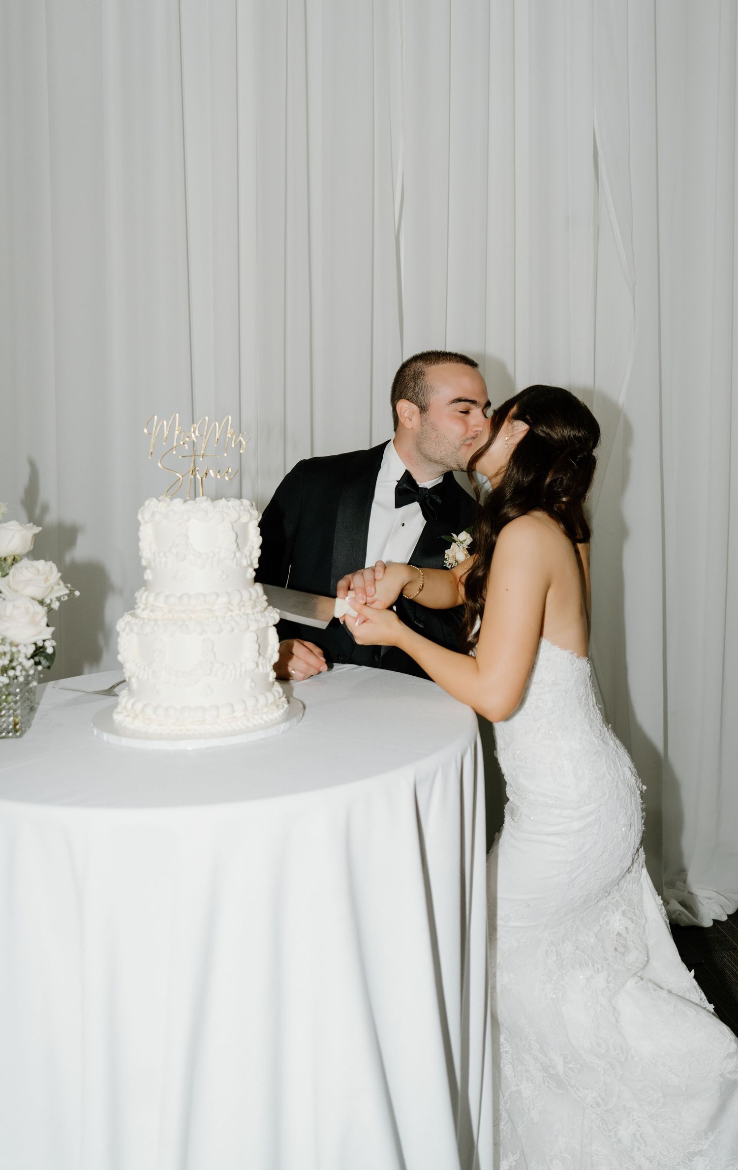A couple shares a romantic kiss beside their elegant white wedding cake at their reception.