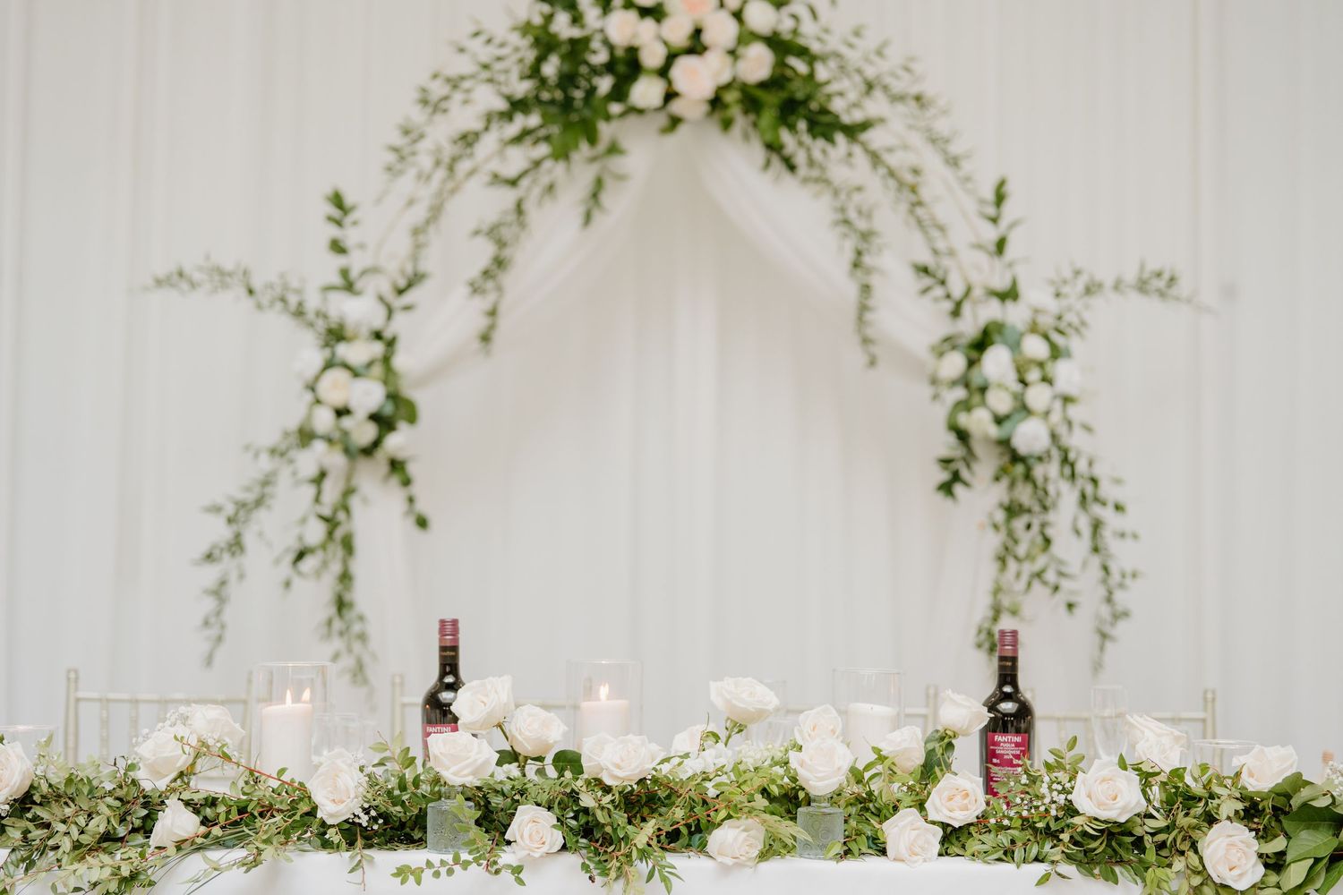 Elegant wedding reception table with hanging greenery, white floral arrangements and glowing candles.