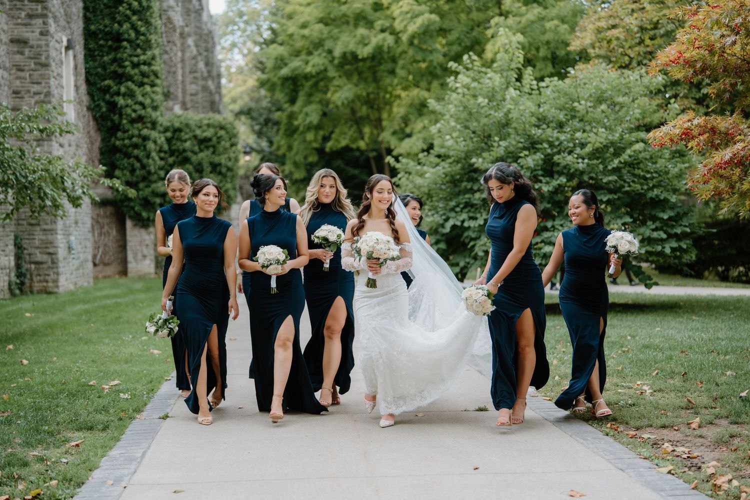 Bridal party in navy blue dresses walks together on garden path while helping bride with flowing white gown.