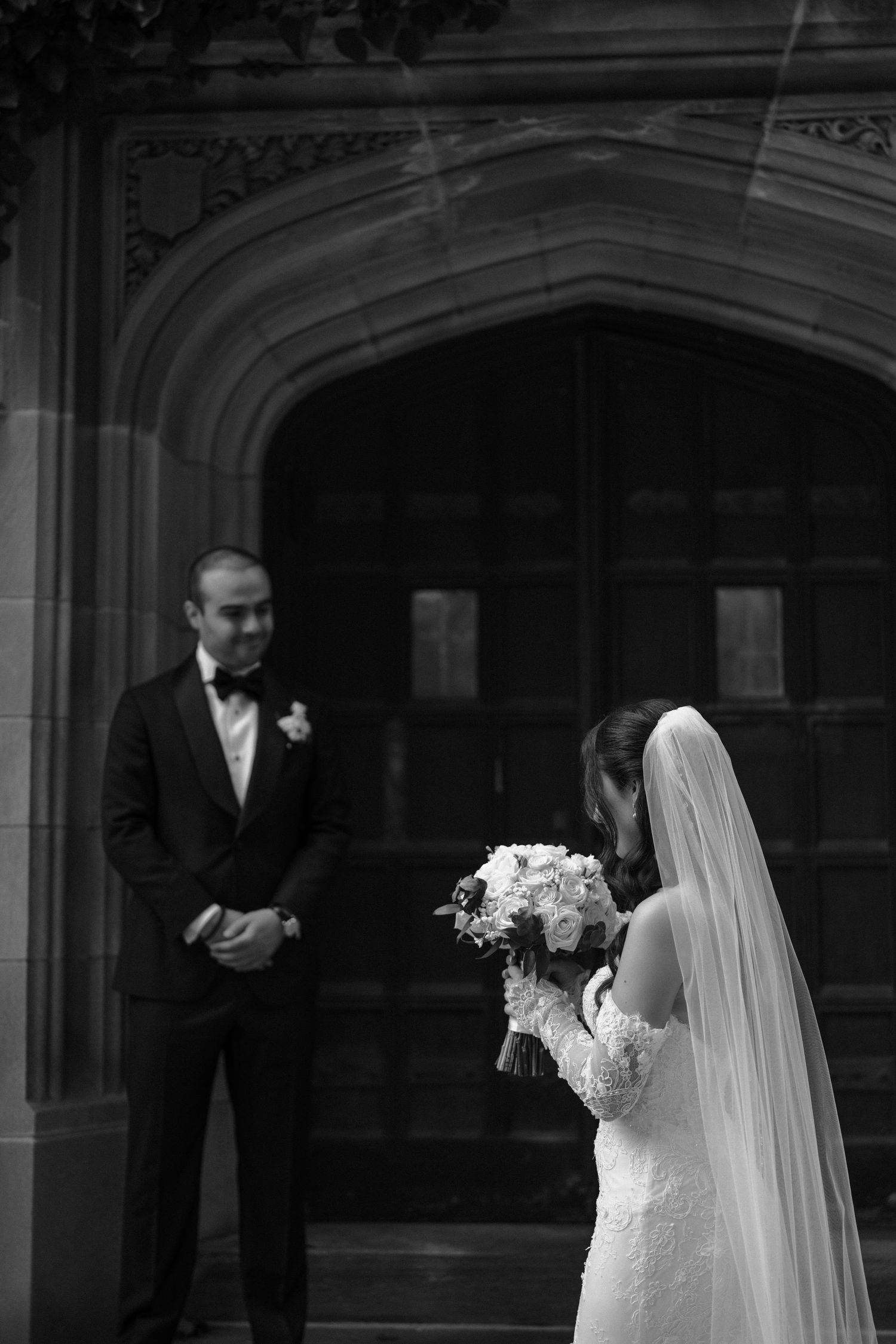 Black and white photo of a bride in a lace wedding dress and veil standing in an ornate archway.