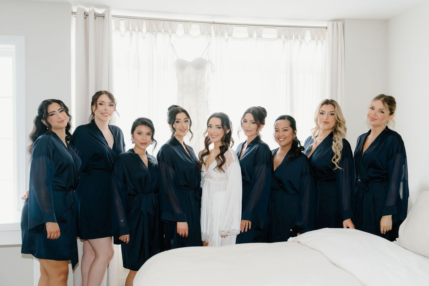 A bridal party wearing matching navy robes poses together in a bright white room before the wedding ceremony.