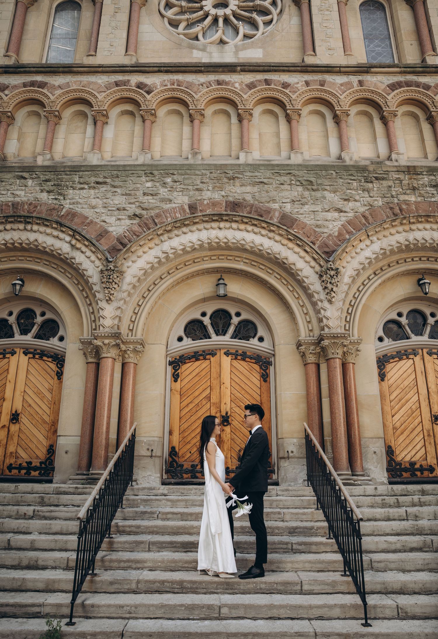 A couple stands on church steps in front of ornate Romanesque arches and wooden doors during a wedding photoshoot.