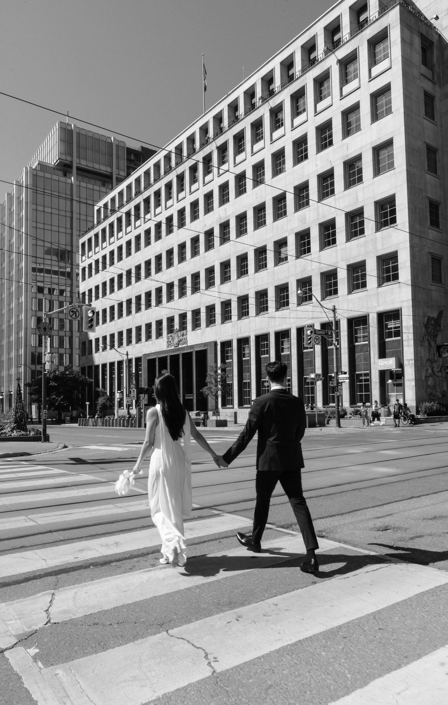 A couple in wedding attire holding hands while crossing a crosswalk in front of a modern office building in black and white.