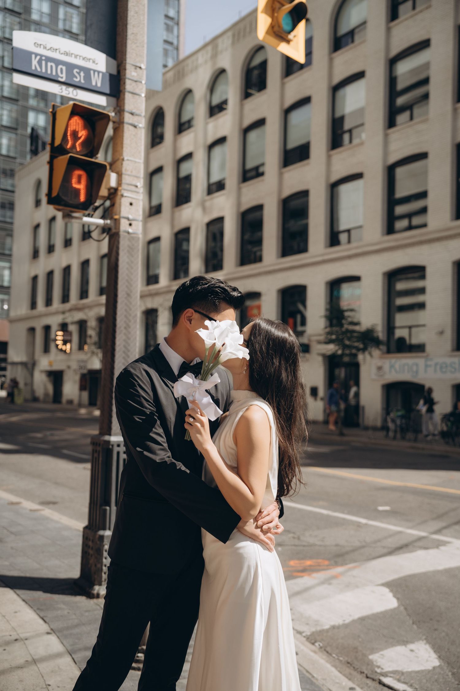A couple in wedding attire embraces on a city street corner with traffic lights and buildings in the background.