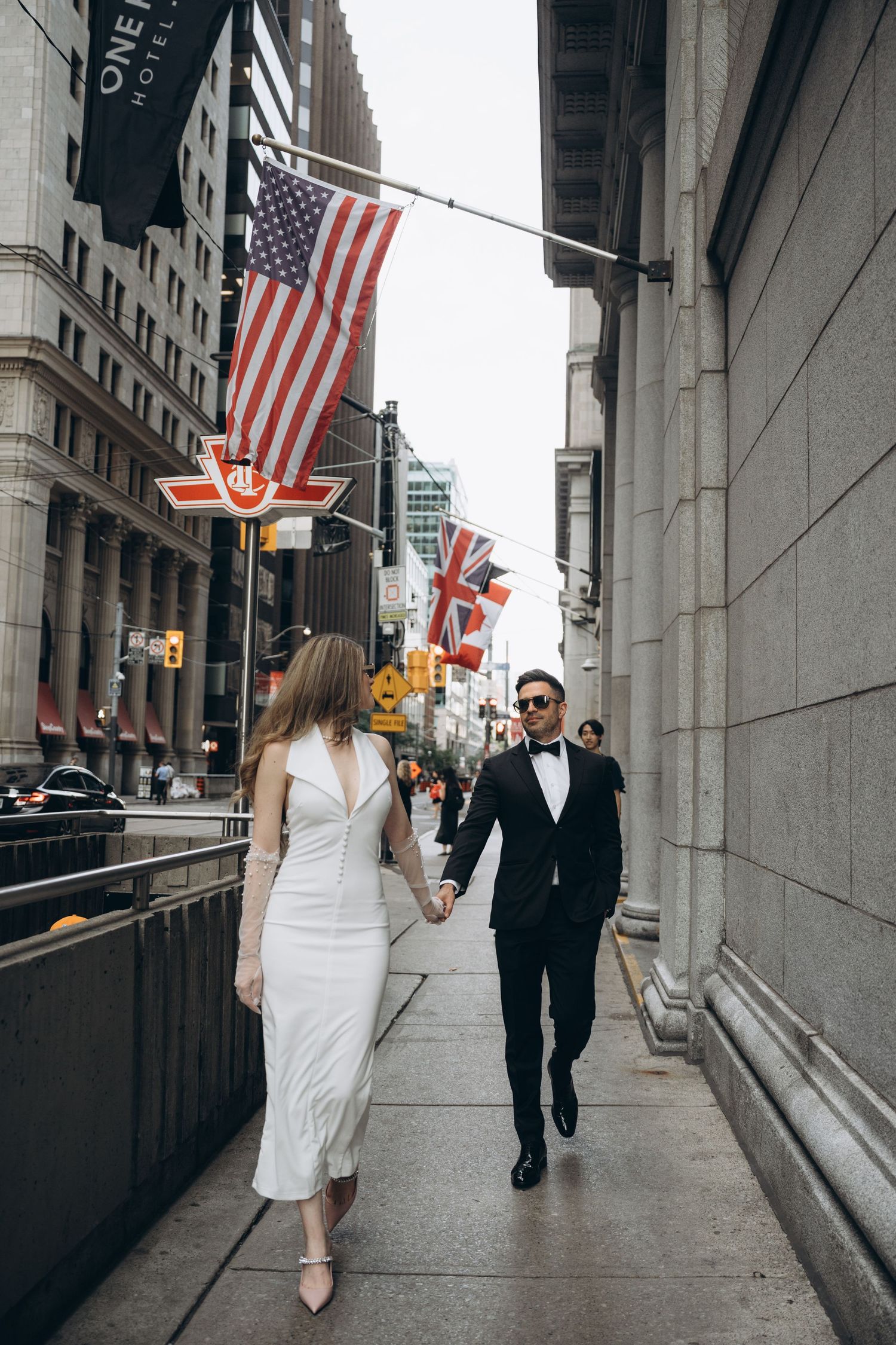 A couple strolls down an urban sidewalk with American flags displayed on the building exterior.