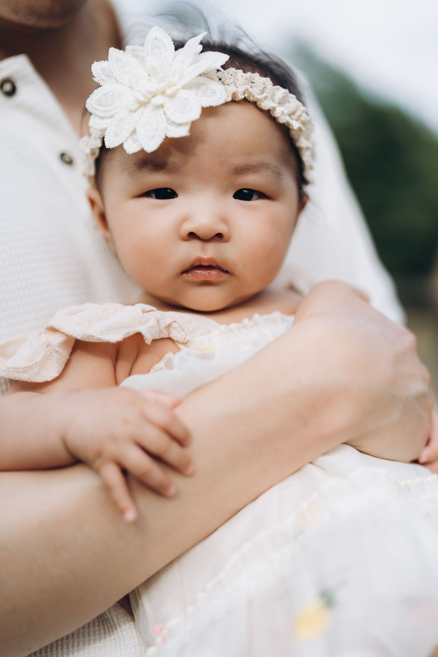 Sweet baby in all white outfit and delicate floral headband lies cradled in gentle arms during photoshoot session.