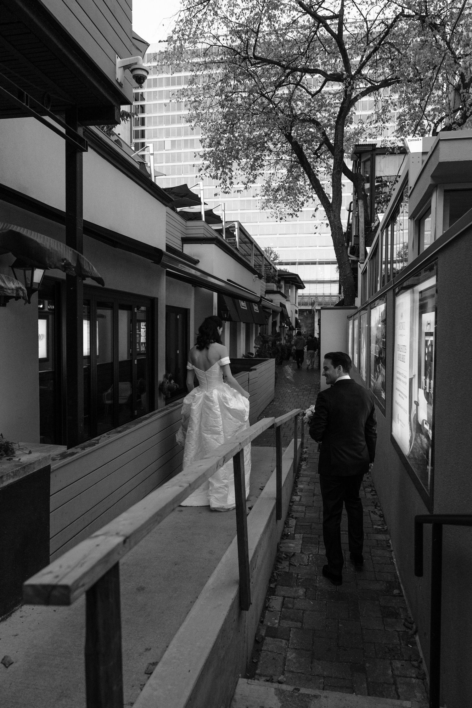 People walking through a covered urban walkway with storefronts and trees visible in black and white photography.