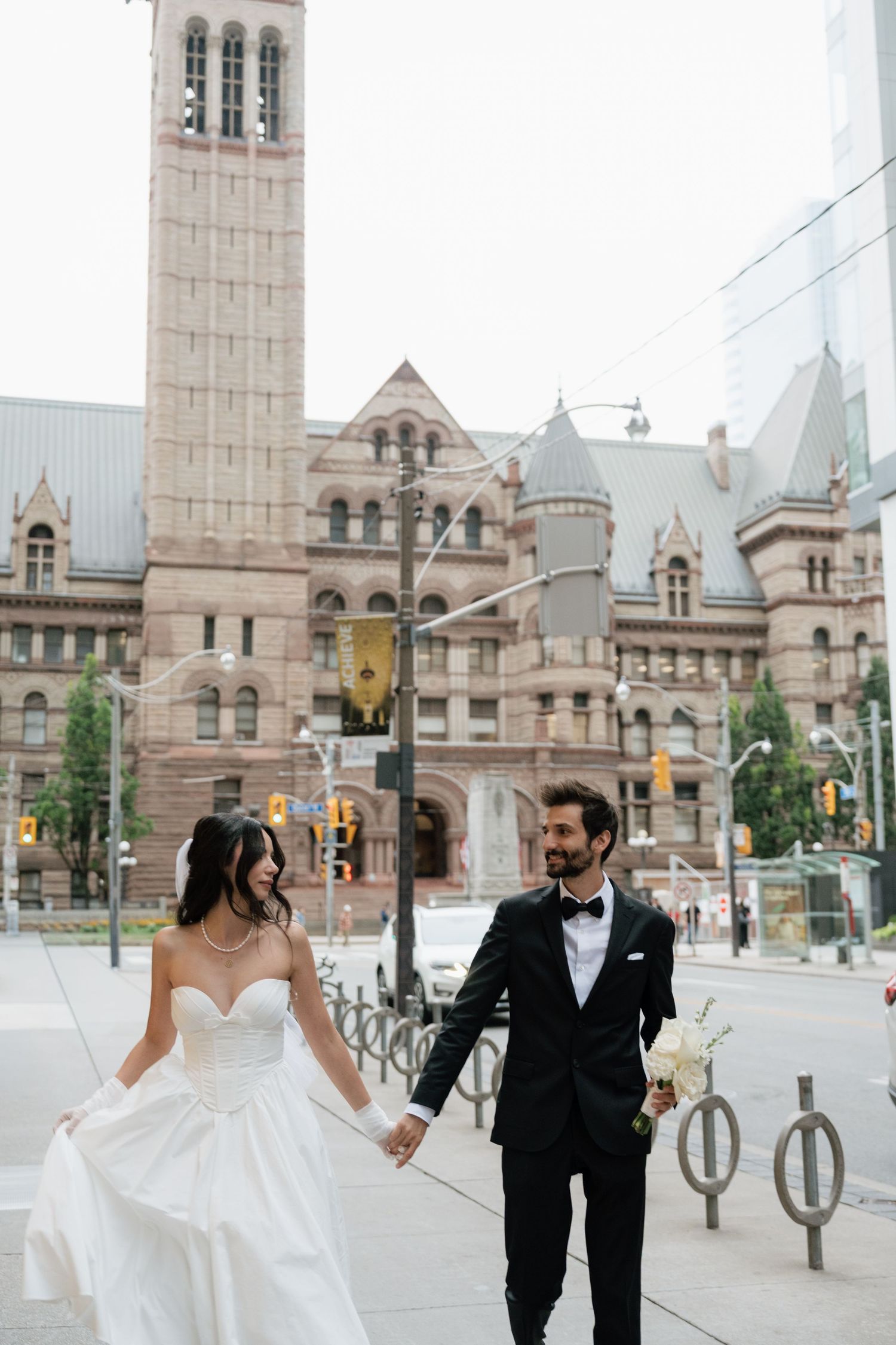 A couple walks hand in hand outside Toronto's Old City Hall, with its Romanesque Revival architecture in the background.