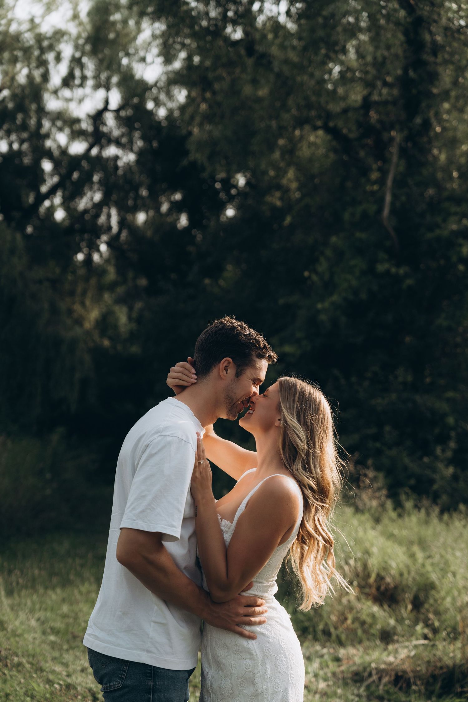 A romantic couple embraces in a sunlit outdoor setting with trees and grass in the background.