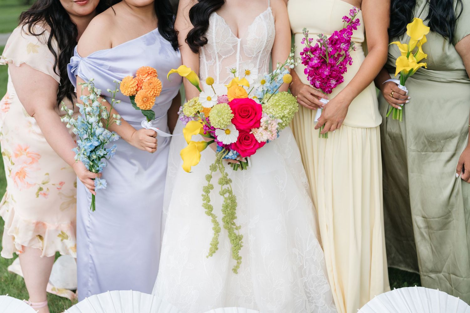 A row of bridesmaids in pastel dresses holding vibrant bouquets with pink, yellow, and purple flowers at an outdoor wedding.