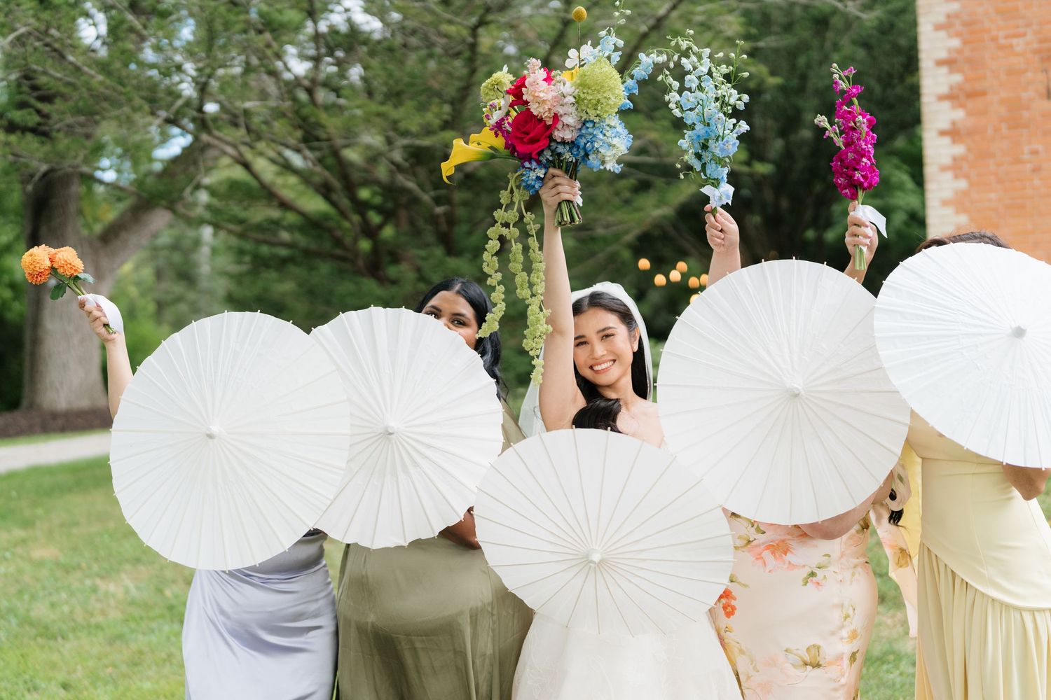 Wedding guests hold white parasols while standing in a row during an outdoor summer celebration.