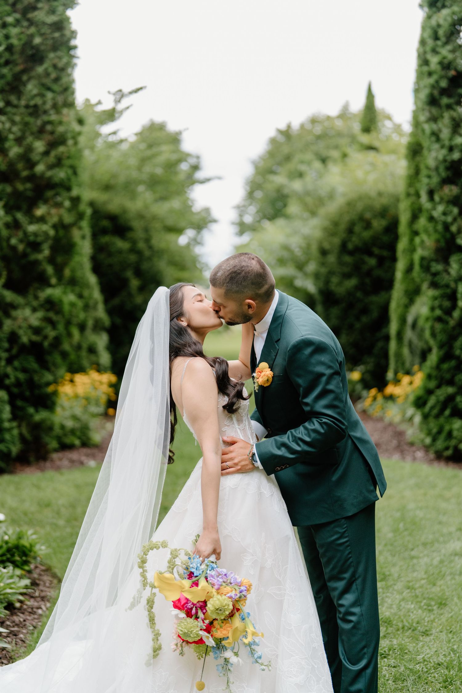 Couple shares romantic kiss in garden setting with bride holding colorful spring bouquet and wearing flowing veil.