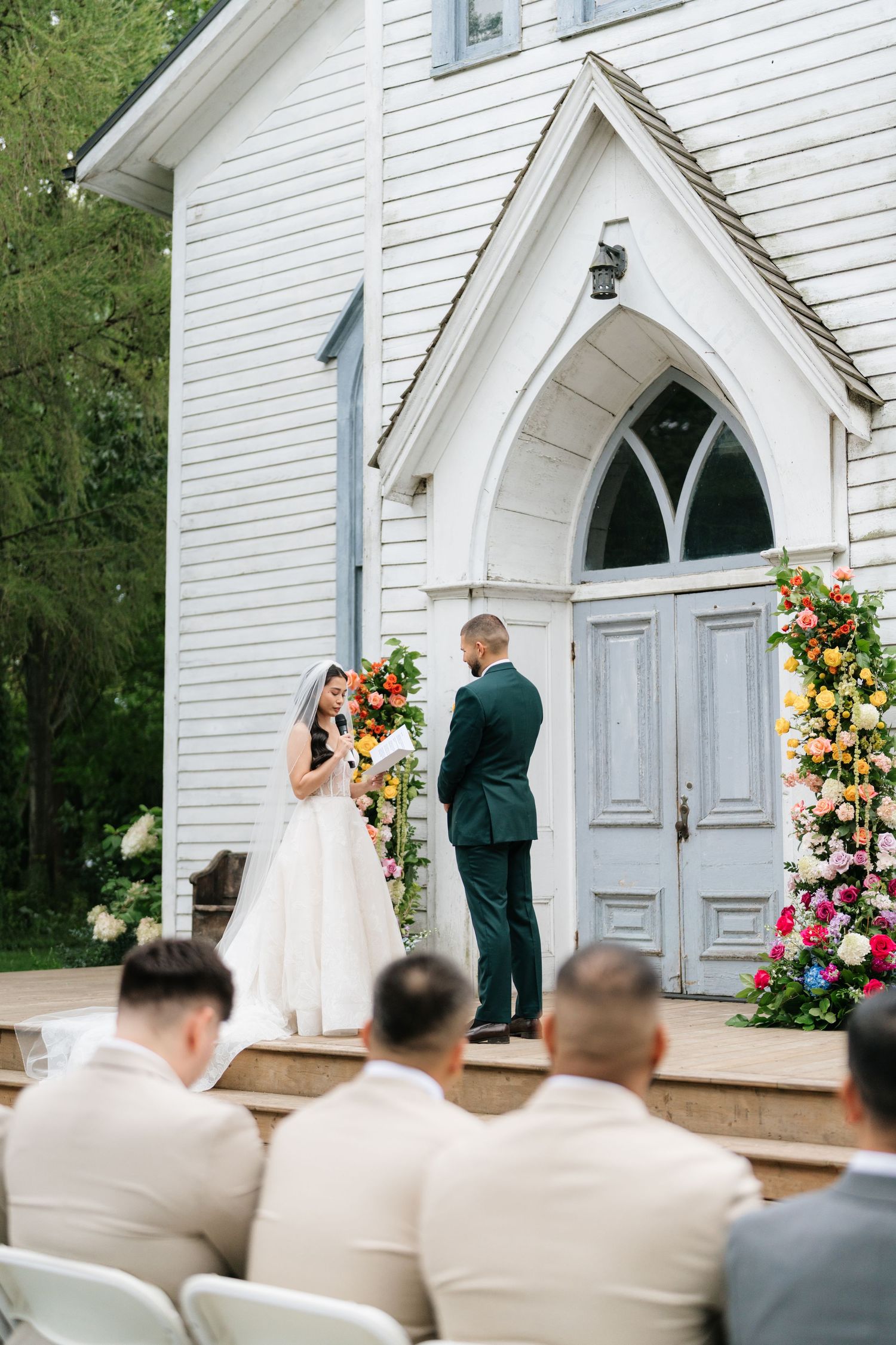 A small white chapel wedding ceremony with floral arrangements and guests seated in beige chairs.