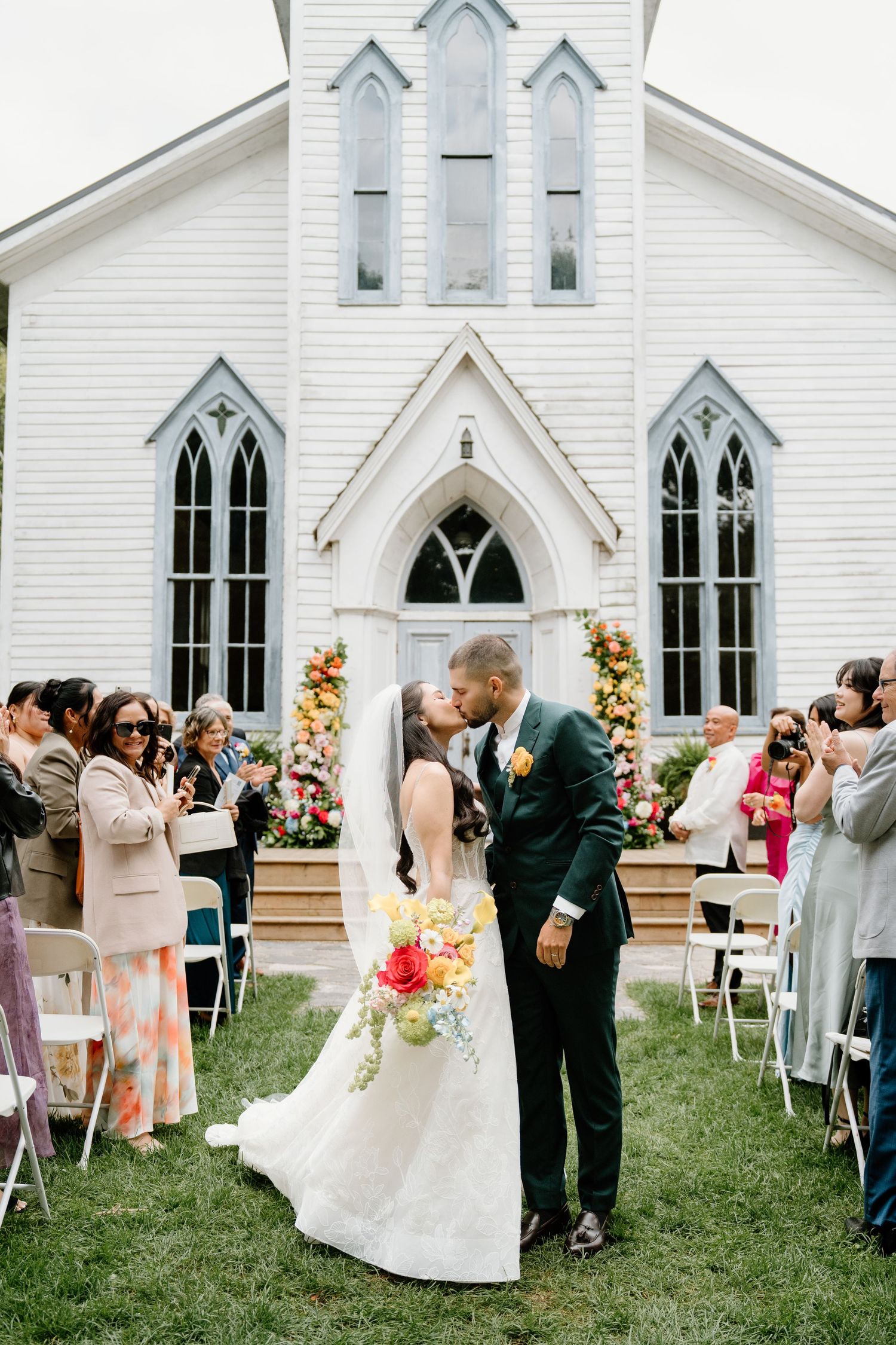 Wedding guests gather on the lawn outside a white church for post-ceremony celebrations.