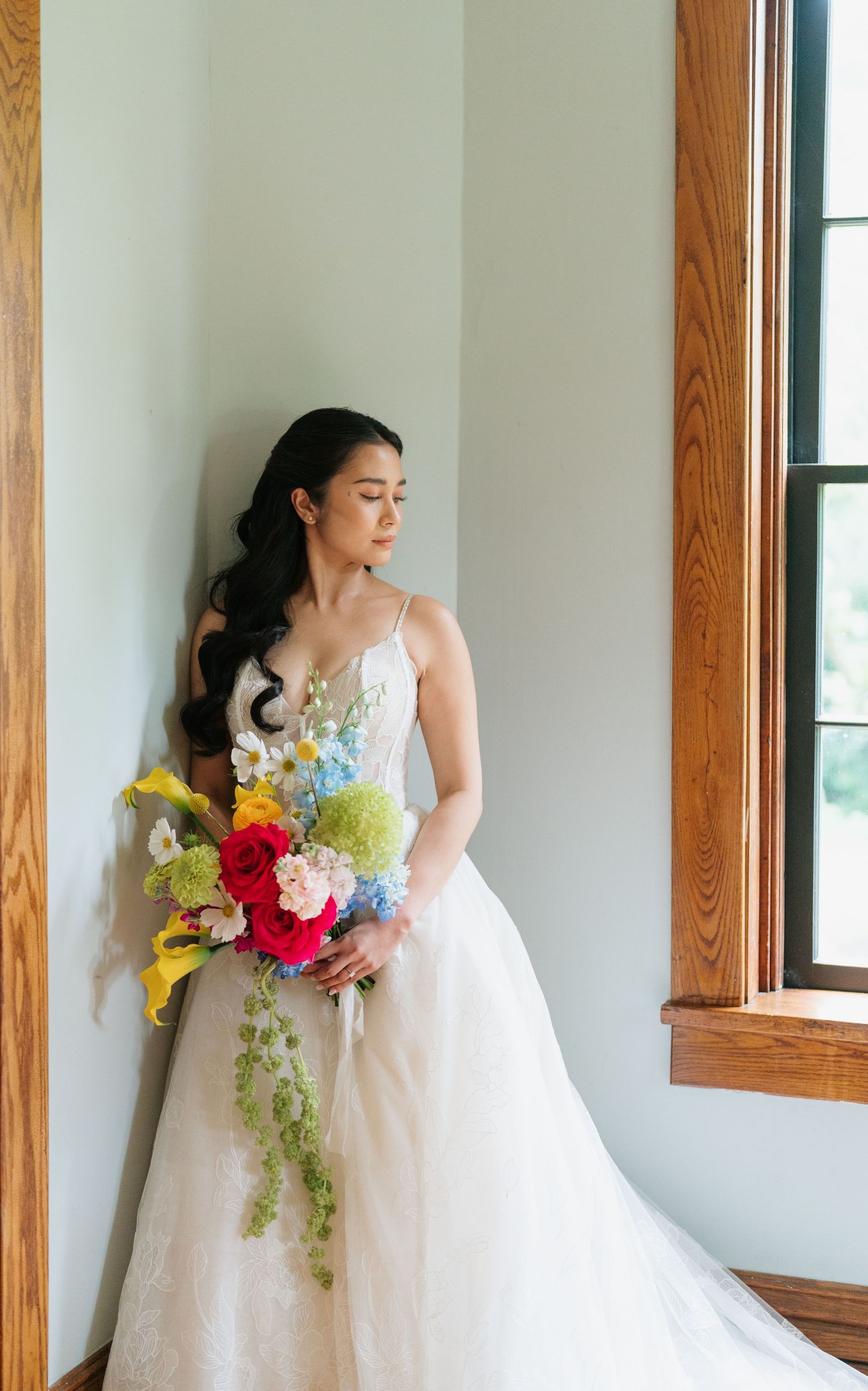 A bride in a white wedding gown holds a vibrant bouquet of red, yellow and blue flowers against a white wall with wood trim.
