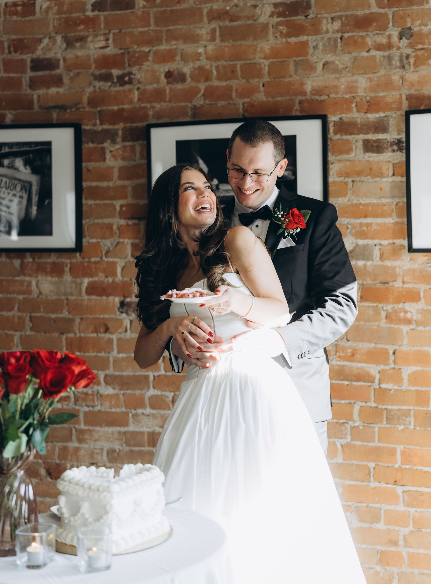 A candid moment during a wedding as newlyweds share laughter while cutting cake against an exposed brick wall.