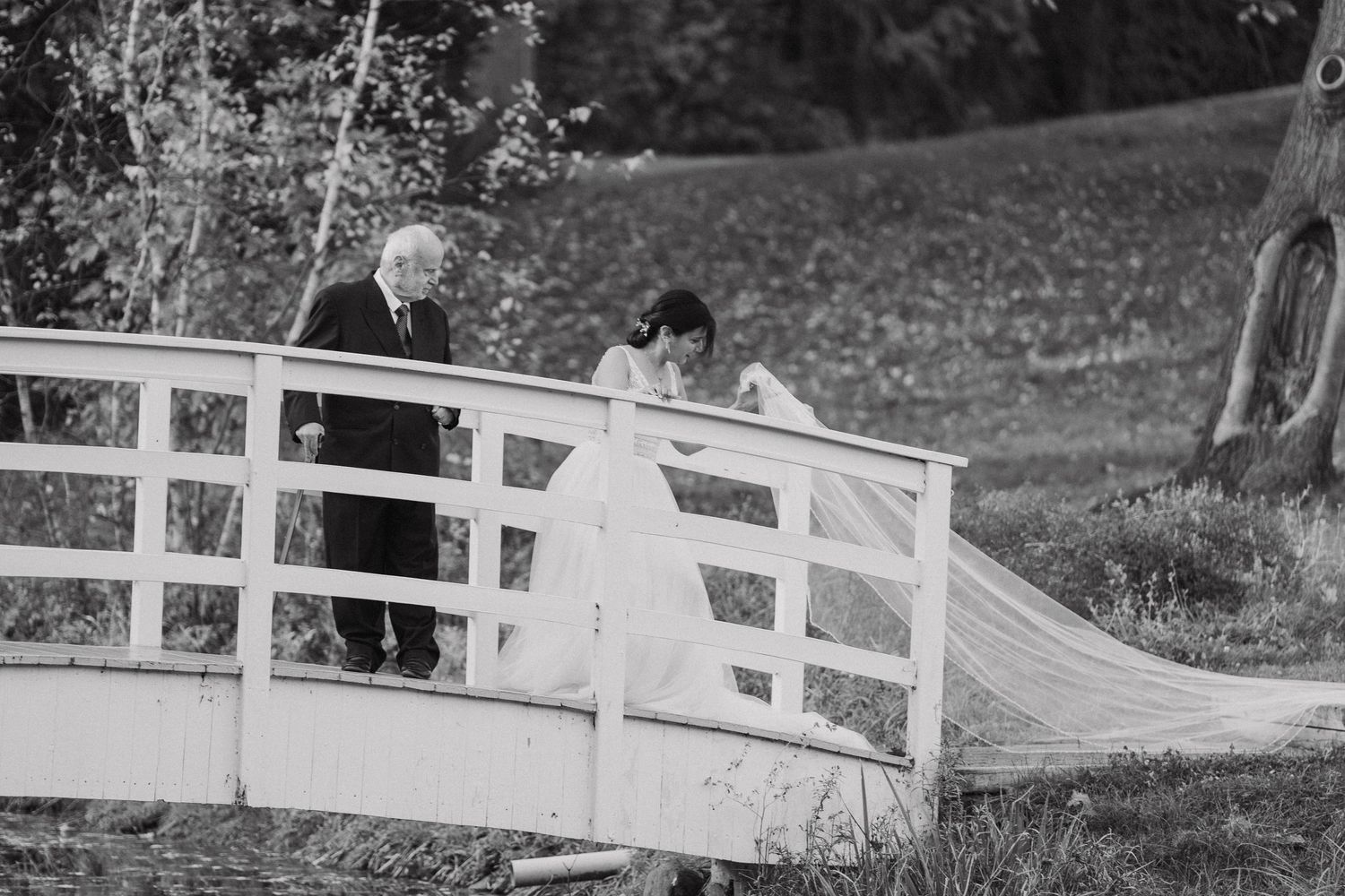 Black and white photo of a wedding couple crossing a white wooden bridge in a garden setting.