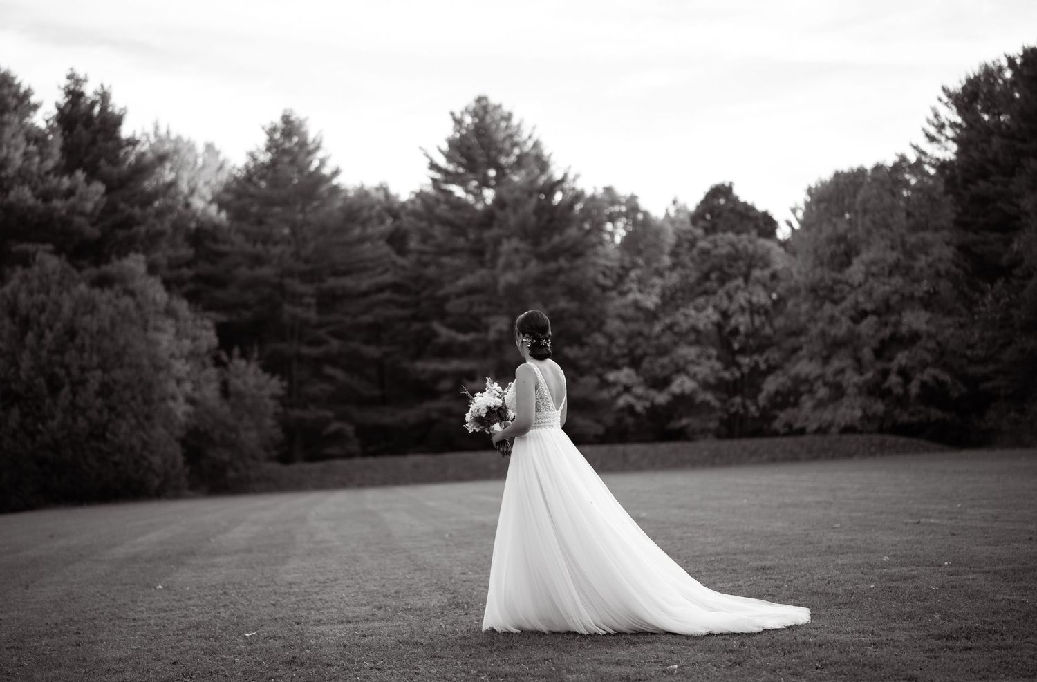 Black and white wedding photo of a bride standing in an open field surrounded by pine trees.
