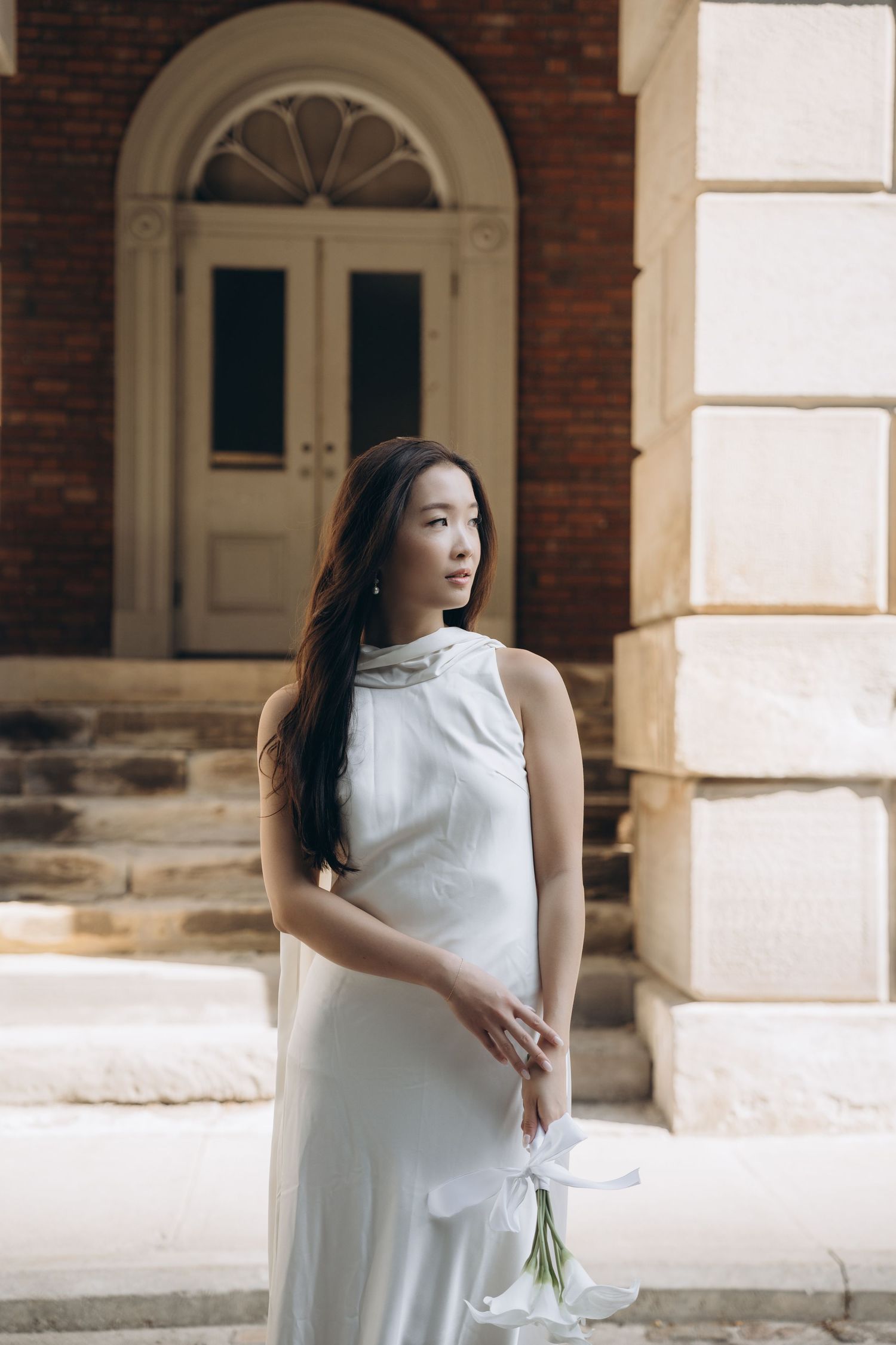 Elegant figure in white sleeveless dress poses outside historic brick building with arched doorway.