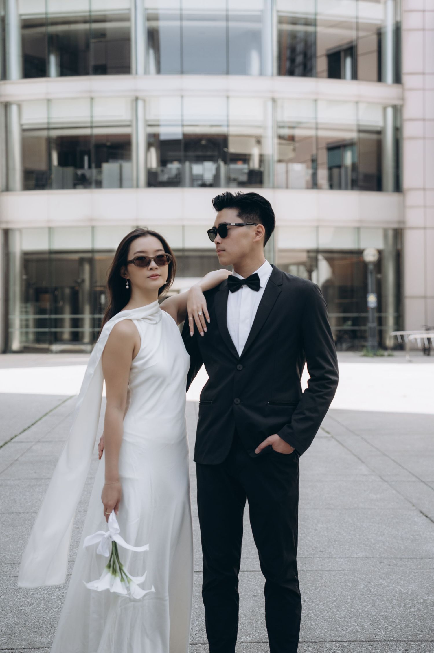 Stylish couple in black and white formal wear with sunglasses stand together against modern architecture.