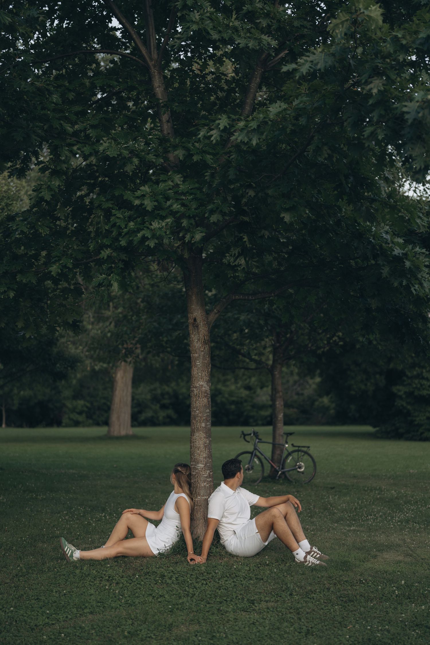 Two people in white outfits sit back-to-back against a tree in a park with a bicycle in the background.