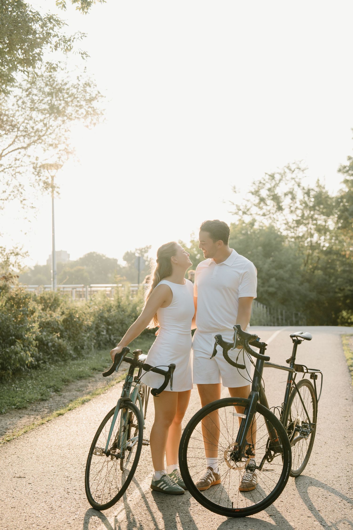 A couple in white outfits share a romantic moment with their bicycles on a sunlit path during golden hour.