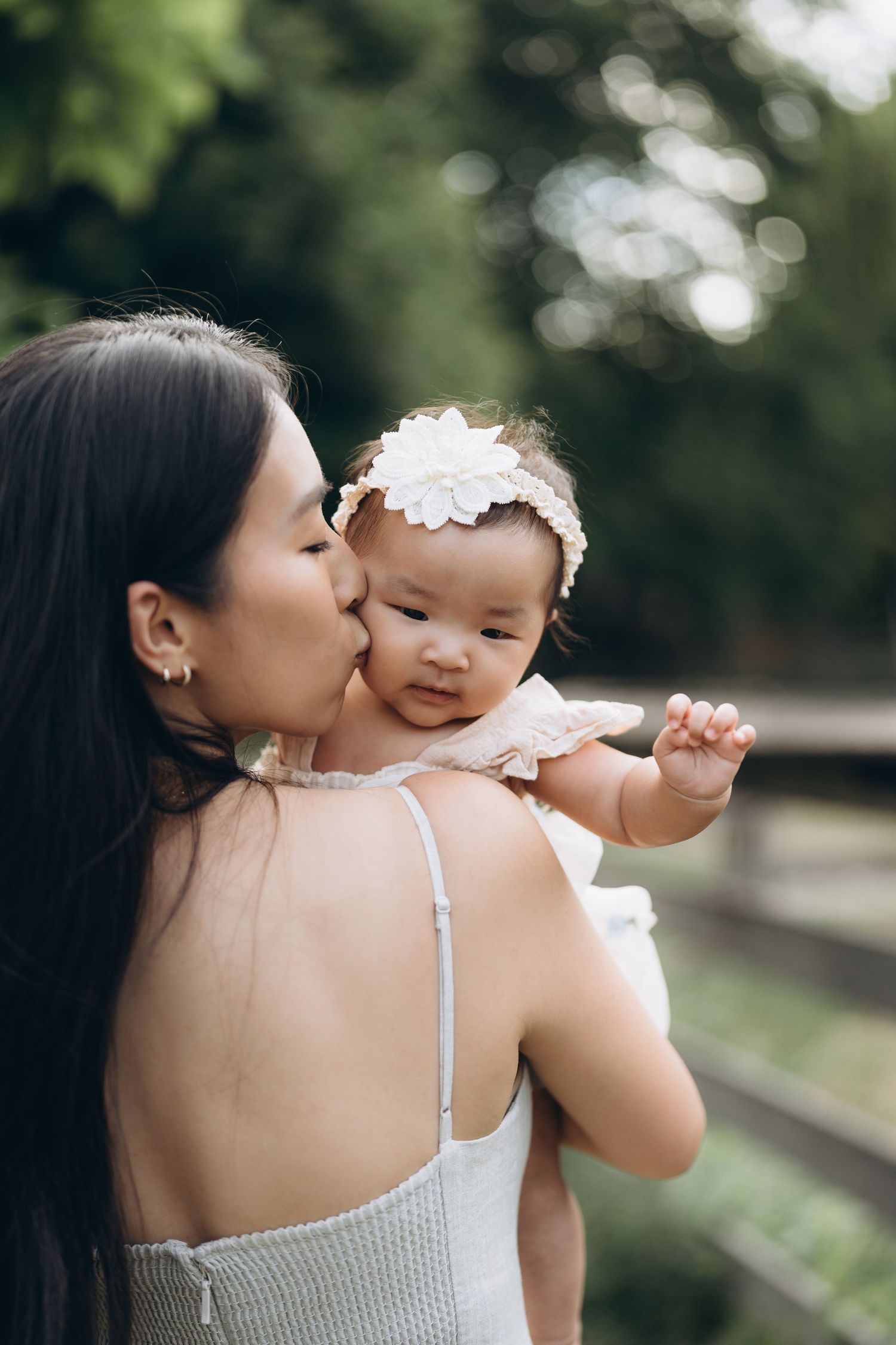 A tender moment between parent and baby in white dresses outdoors with natural bokeh background.