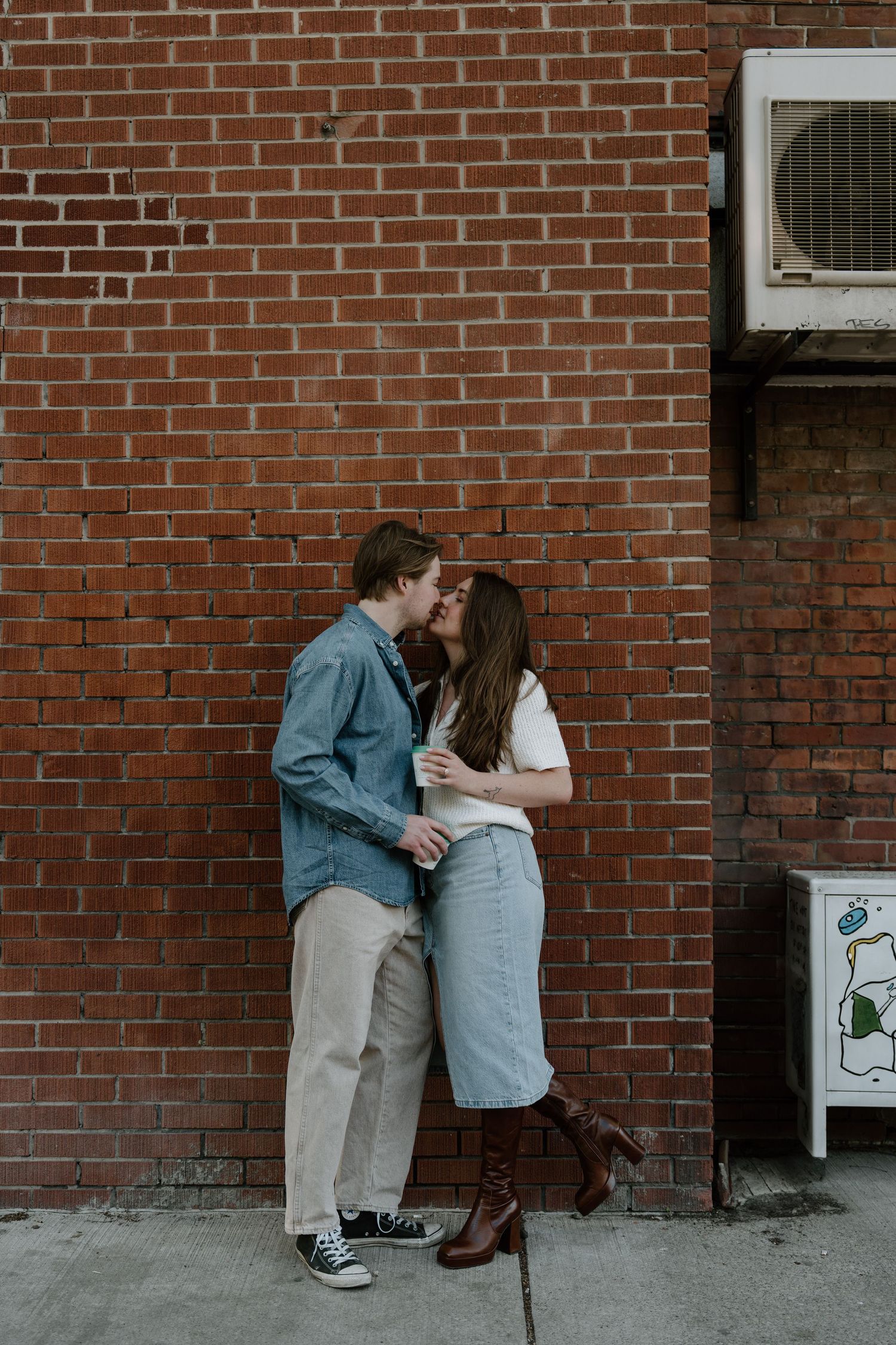 A couple shares an intimate moment against a brick wall in casual outfits on an urban street.