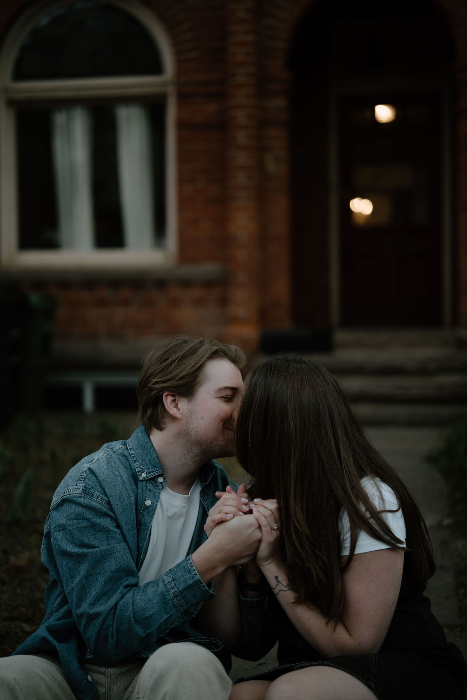 A romantic couple sitting outside a brick building at dusk, holding hands intimately in moody lighting.