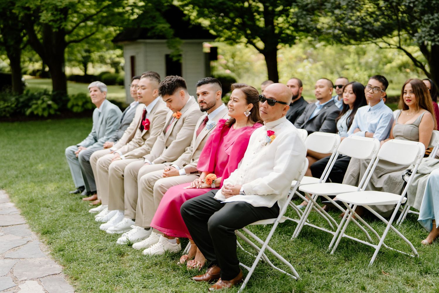 Wedding guests seated in white folding chairs on a grassy lawn during an outdoor ceremony.
