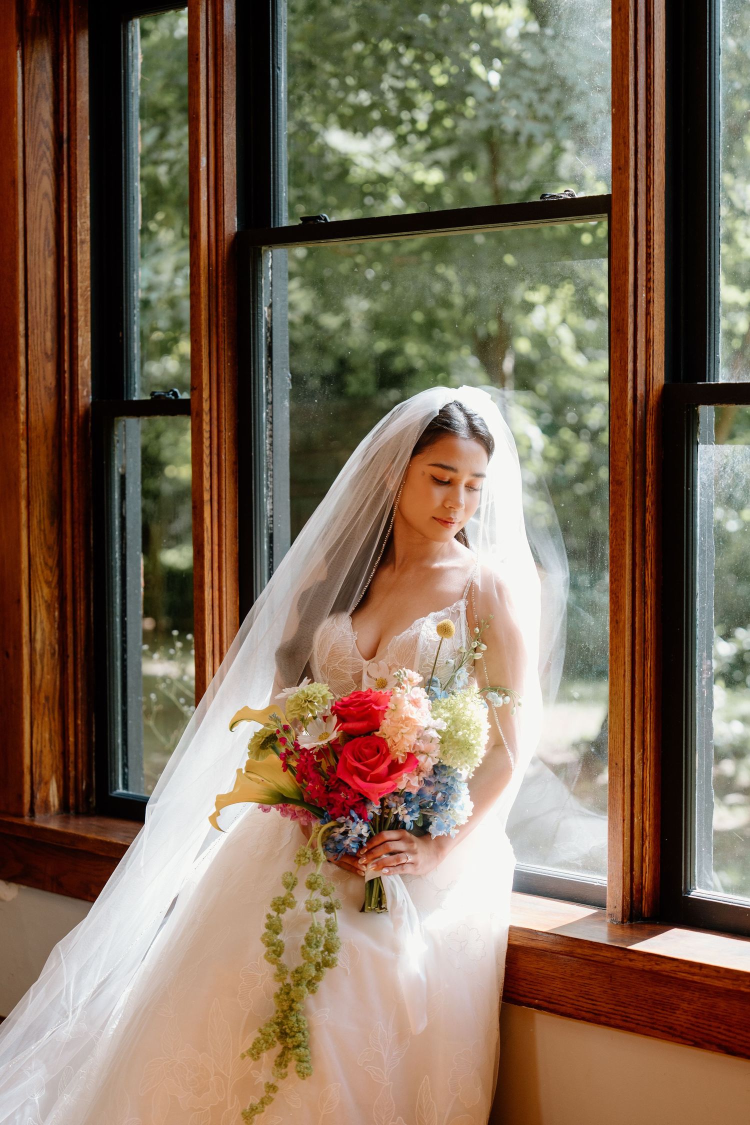 A bride in a flowing white gown and veil poses by sunlit windows while holding a vibrant red and blue bouquet.