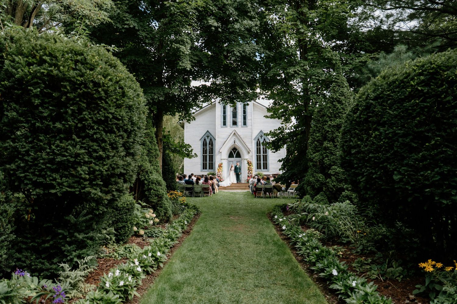 A scenic garden path with blooming flowers leads to a charming white chapel in the distance.