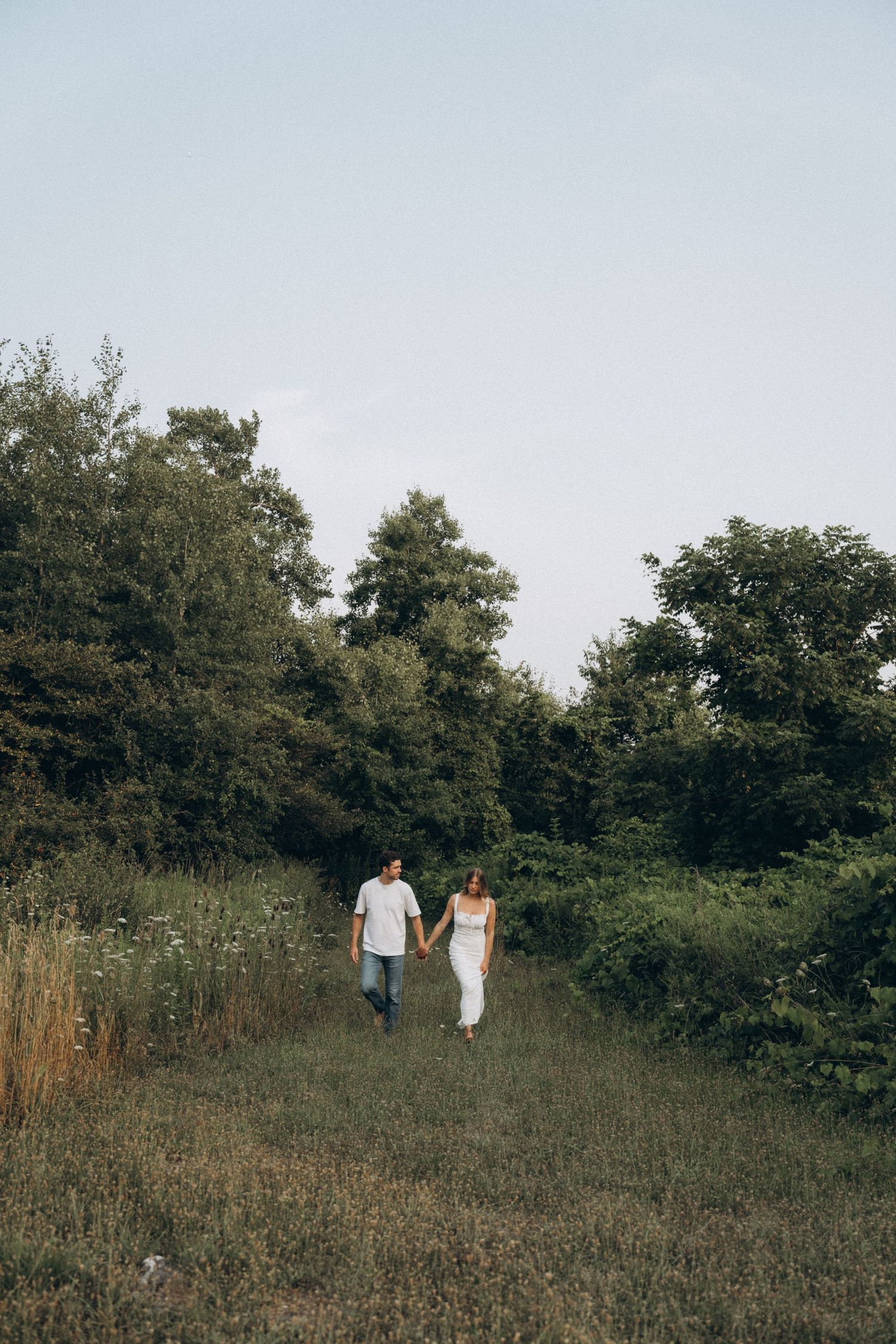 A couple walks hand in hand along a grassy path bordered by trees and wildflowers on a summer day.