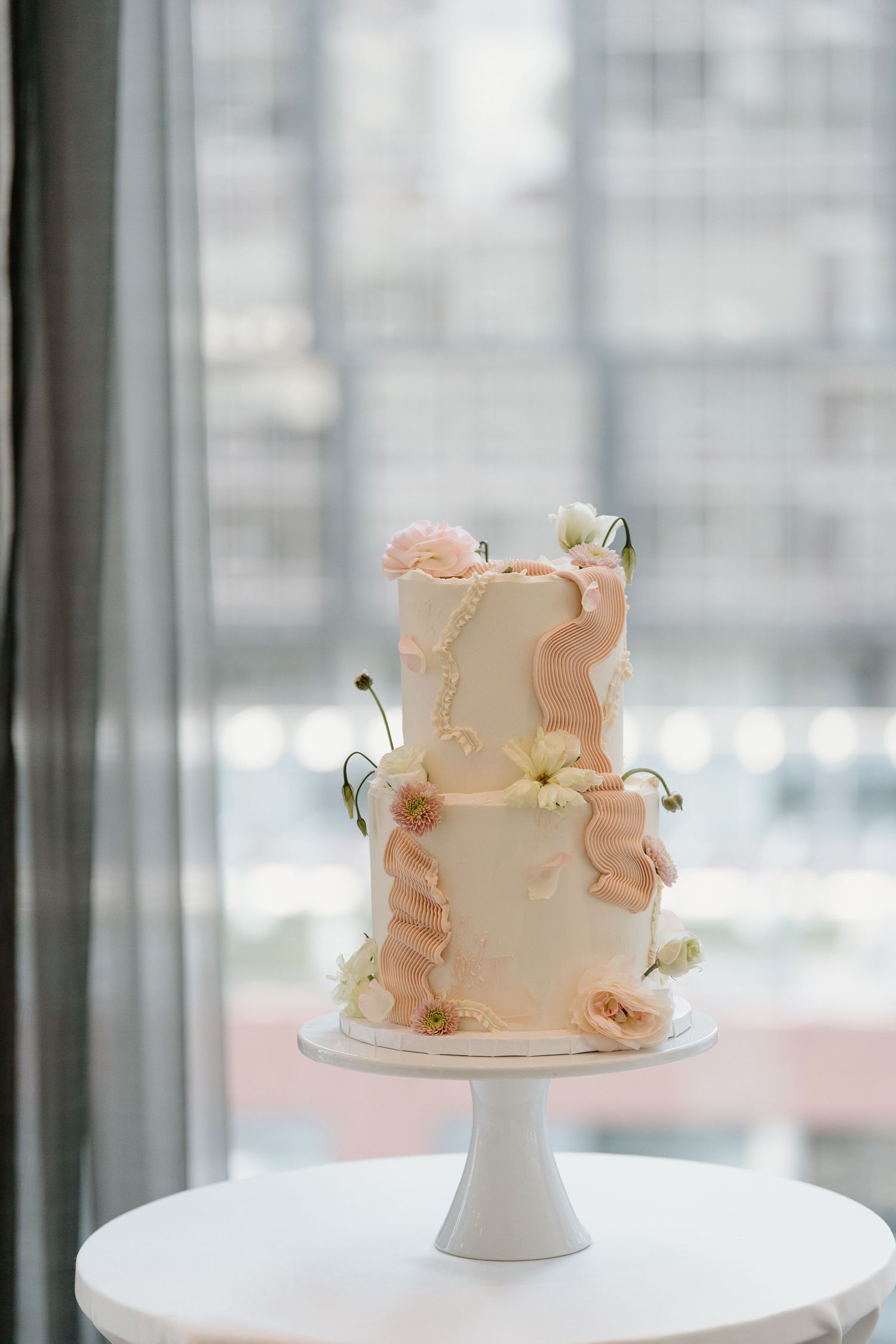 Elegant two-tier white wedding cake with delicate peach ruffles and fresh cherries on a white pedestal stand.
