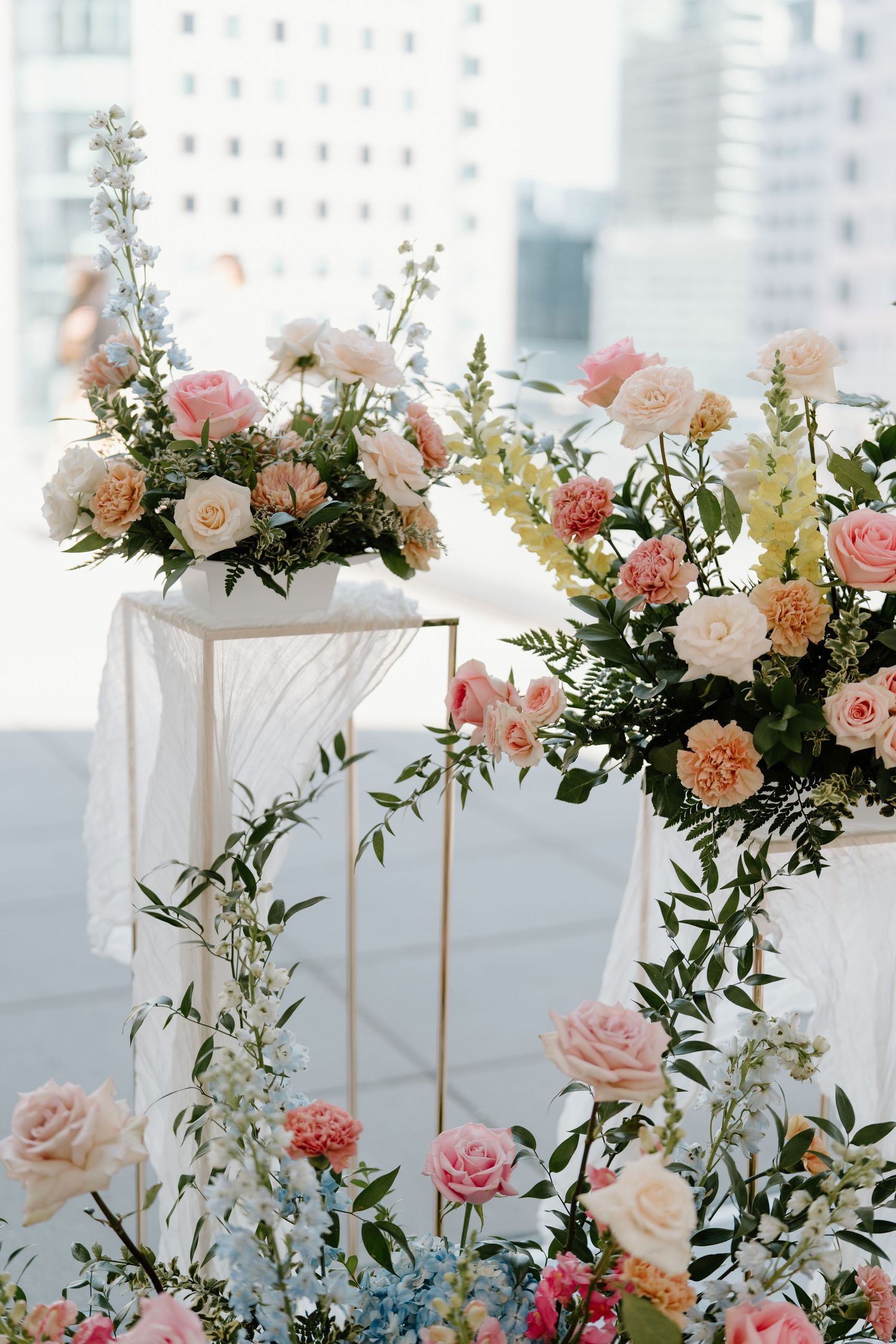 Elegant floral arrangements with pink and white roses on gold pedestals against a city backdrop.