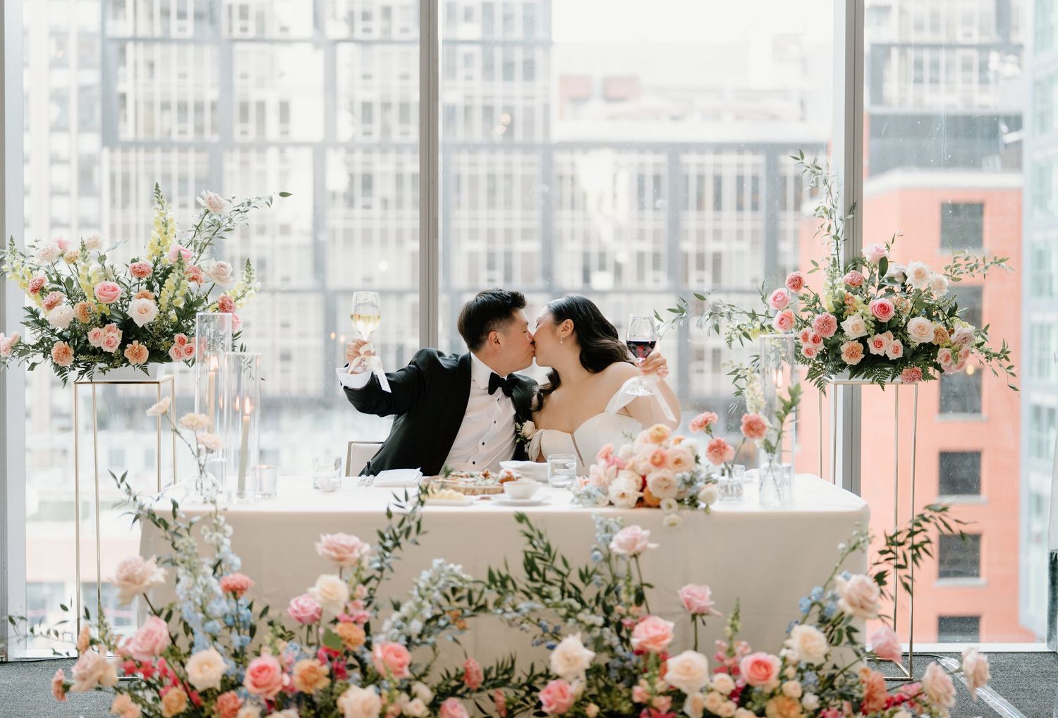 A romantic couple sharing a kiss at an elegant wedding reception with pink floral arrangements and city views through windows.