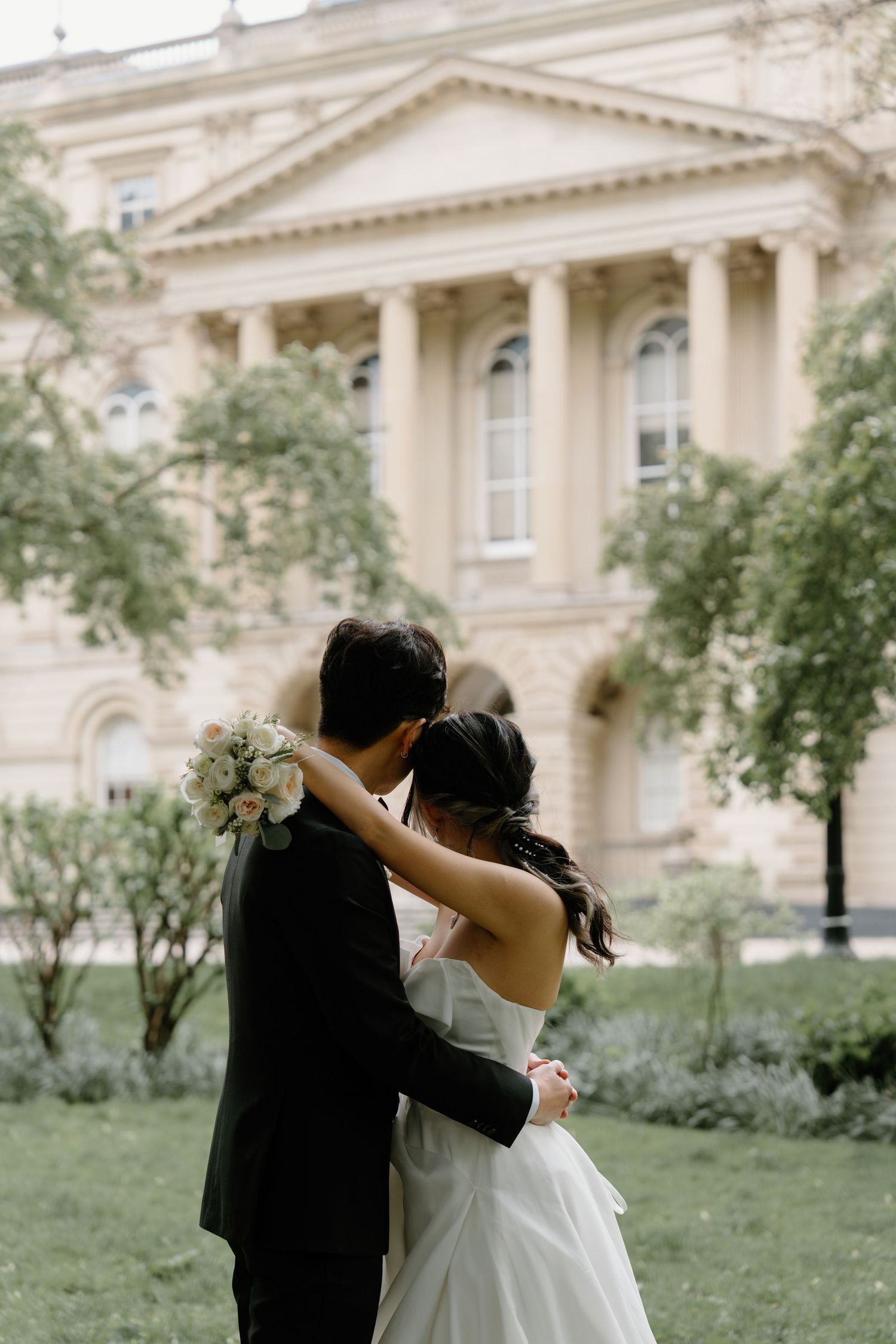A couple embraces in front of a neoclassical building with white columns and arched windows.