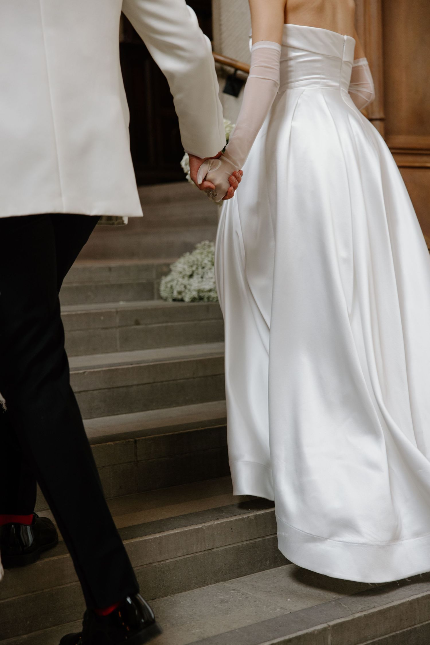 Wedding couple walking up stone steps with focus on bride's white satin gown and groom's black tuxedo shoes.