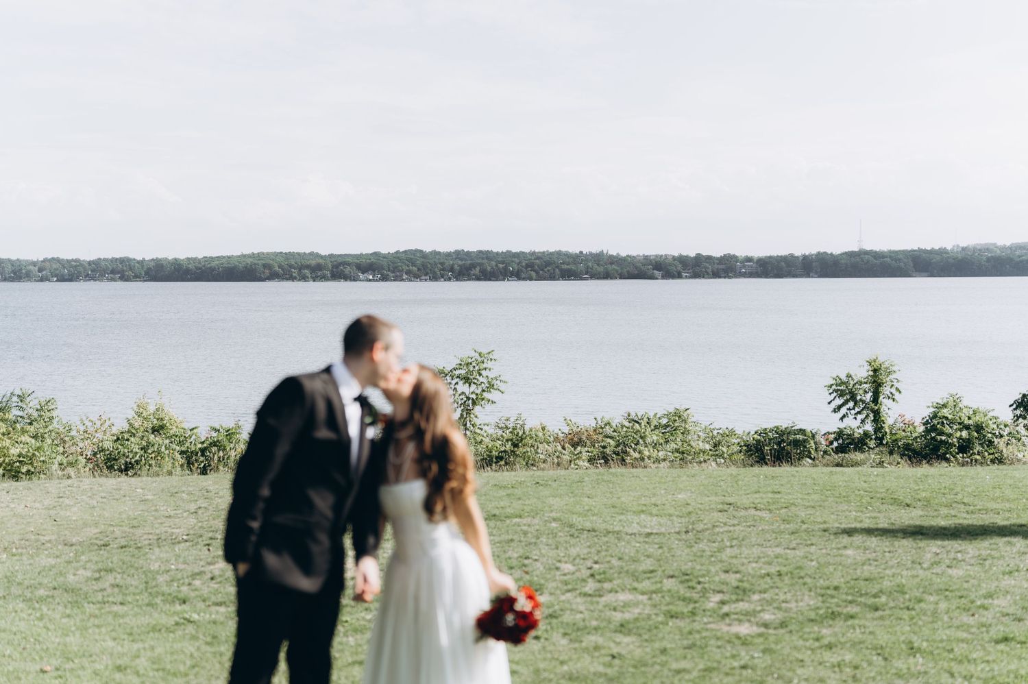 A couple in wedding attire embraces at an outdoor waterfront venue with scenic lake and trees in the background.